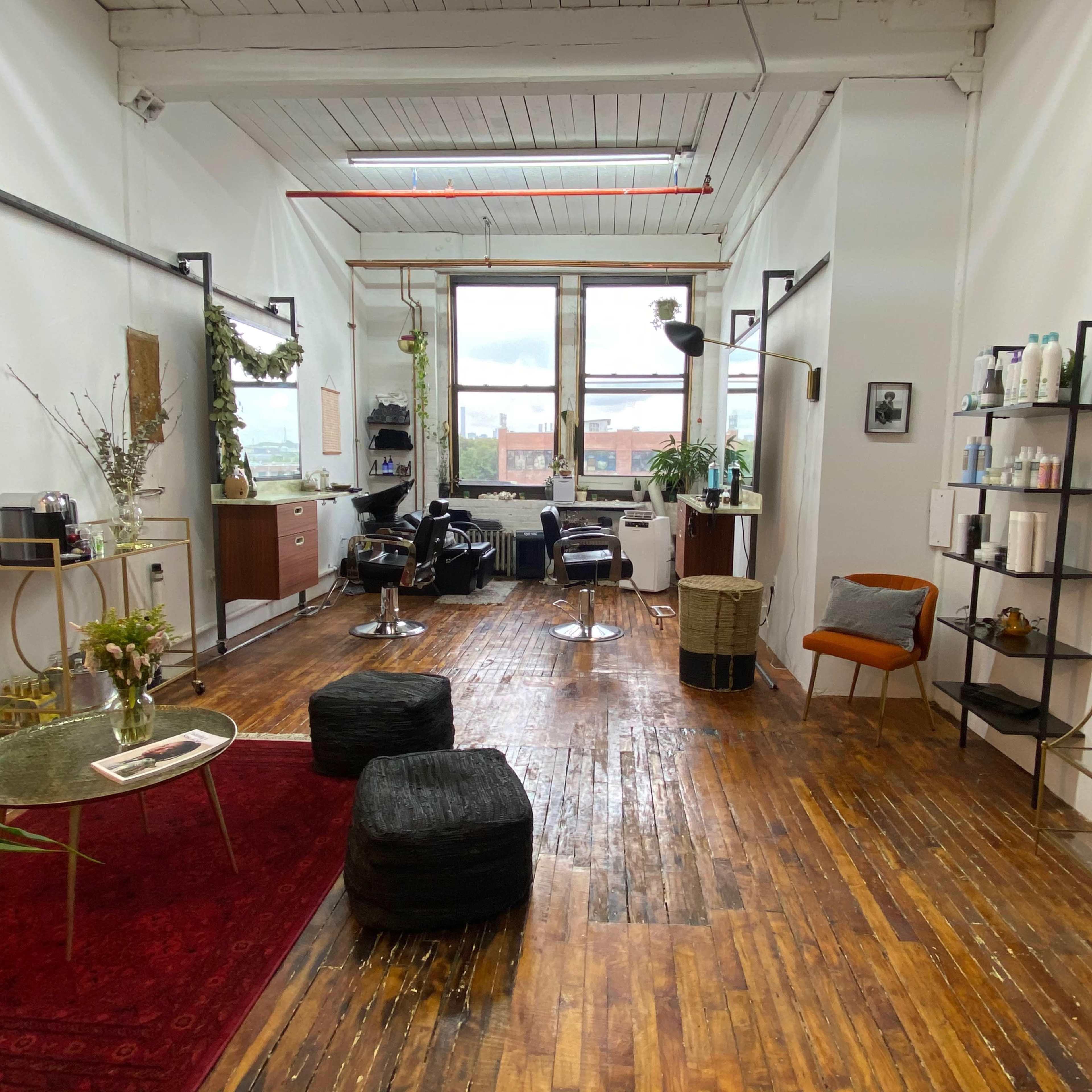 A spacious hair salon with wooden flooring, featuring styling chairs, a mirror, and various hair products arranged on shelves.
