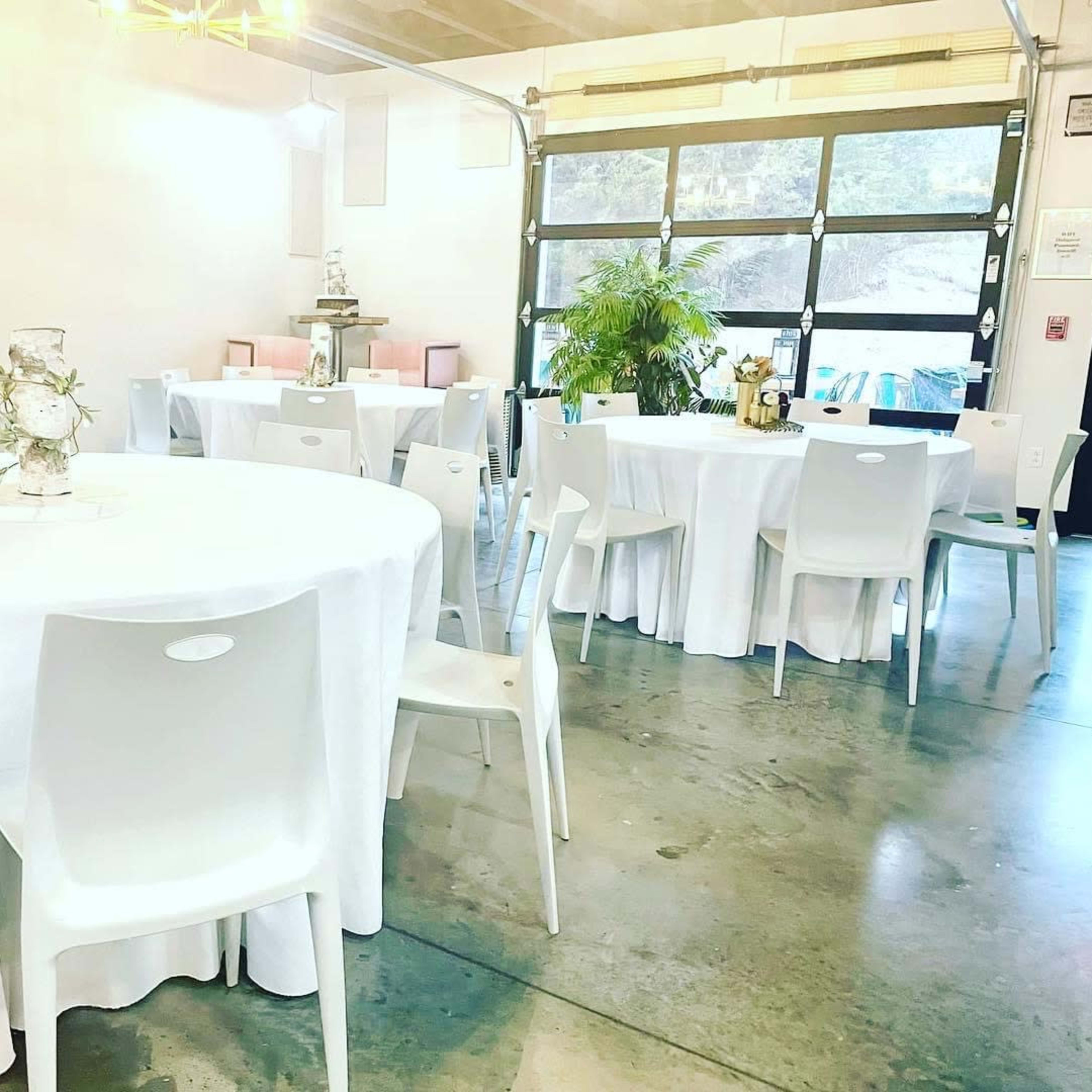 A spacious room with several round tables covered in white tablecloths and white chairs arranged around them, along with a large garage door.