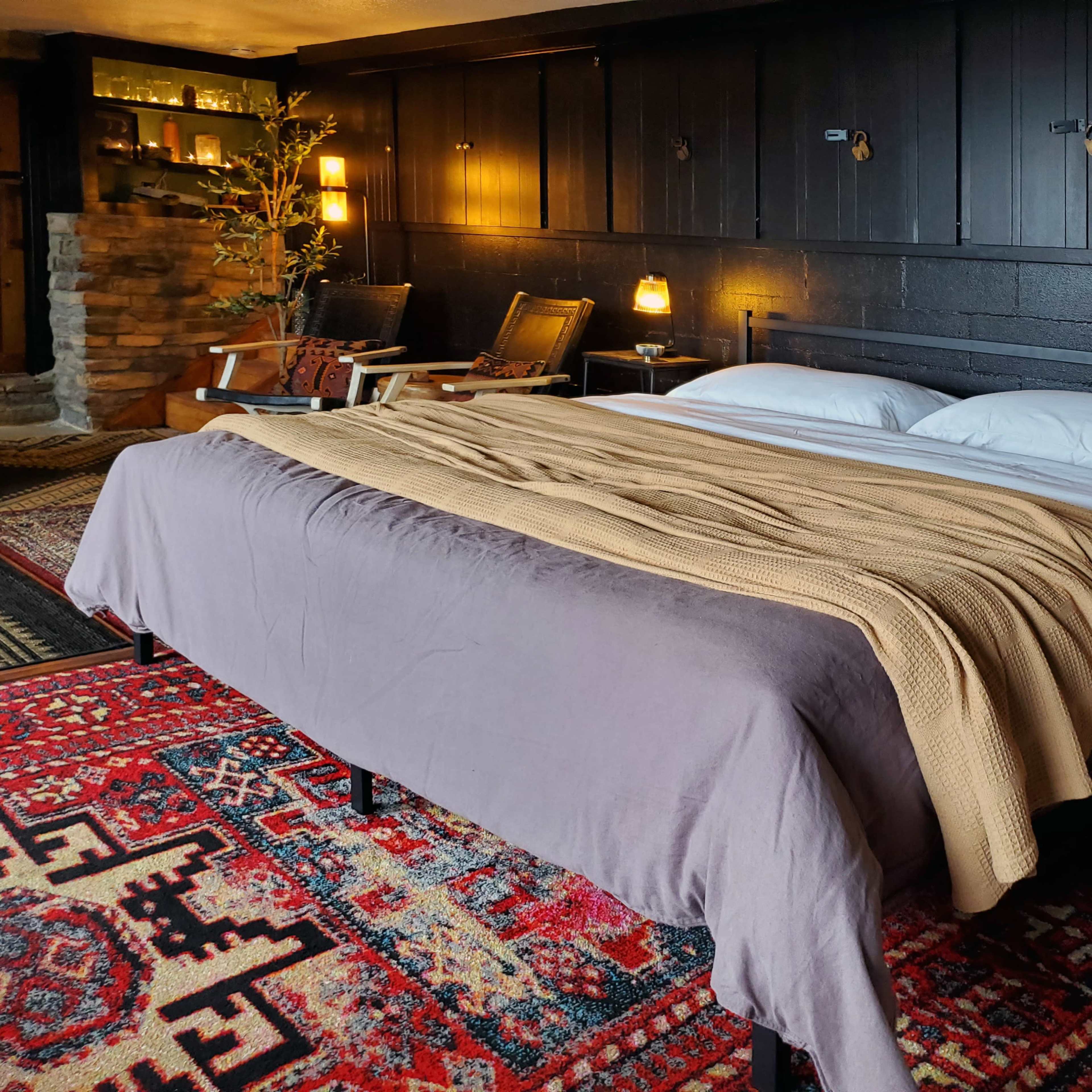 A cozy bedroom with a large bed covered in a beige blanket and two white pillows, surrounded by wooden walls and a patterned rug.