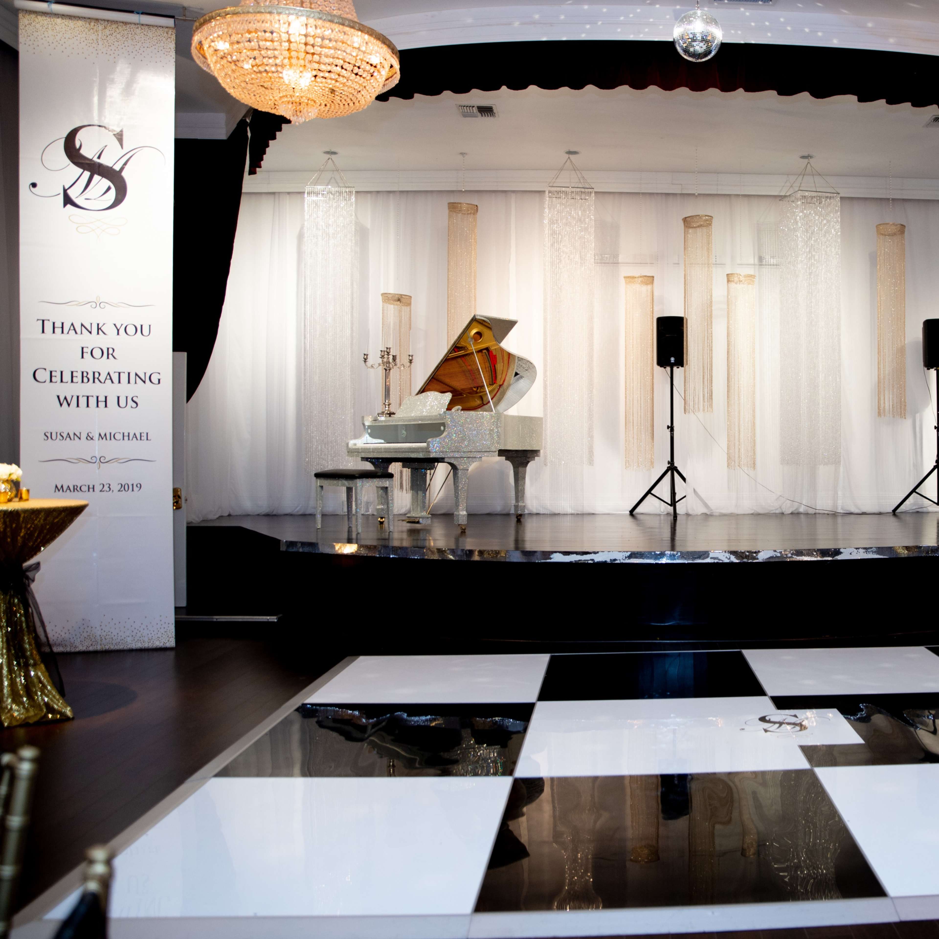A elegantly decorated event space featuring a grand piano on a stage, black and white dance floor tiles, and large banners thanking guests for their attendance.
