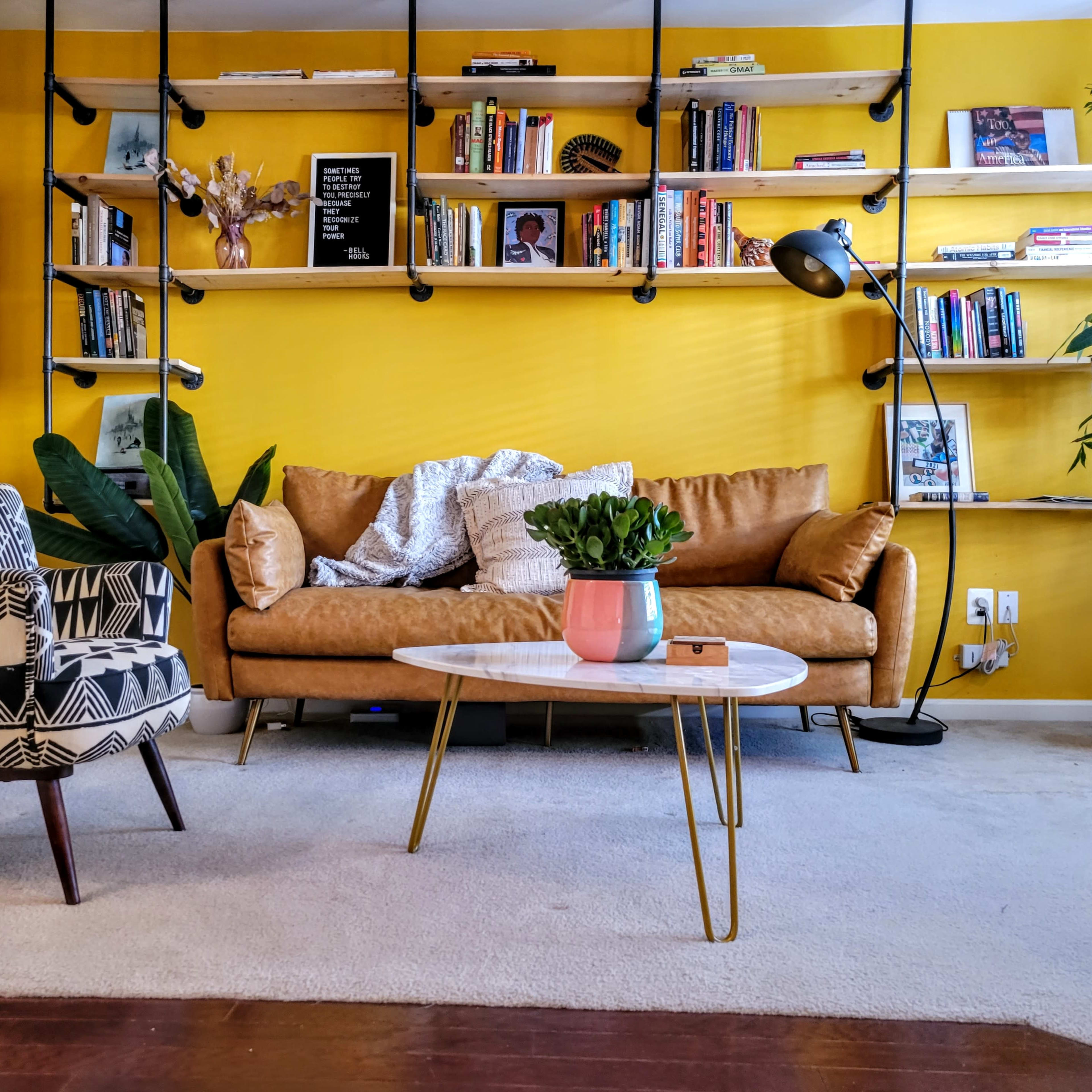 The living room features a yellow wall with bookshelves, a brown leather couch, a round coffee table, and various plants.