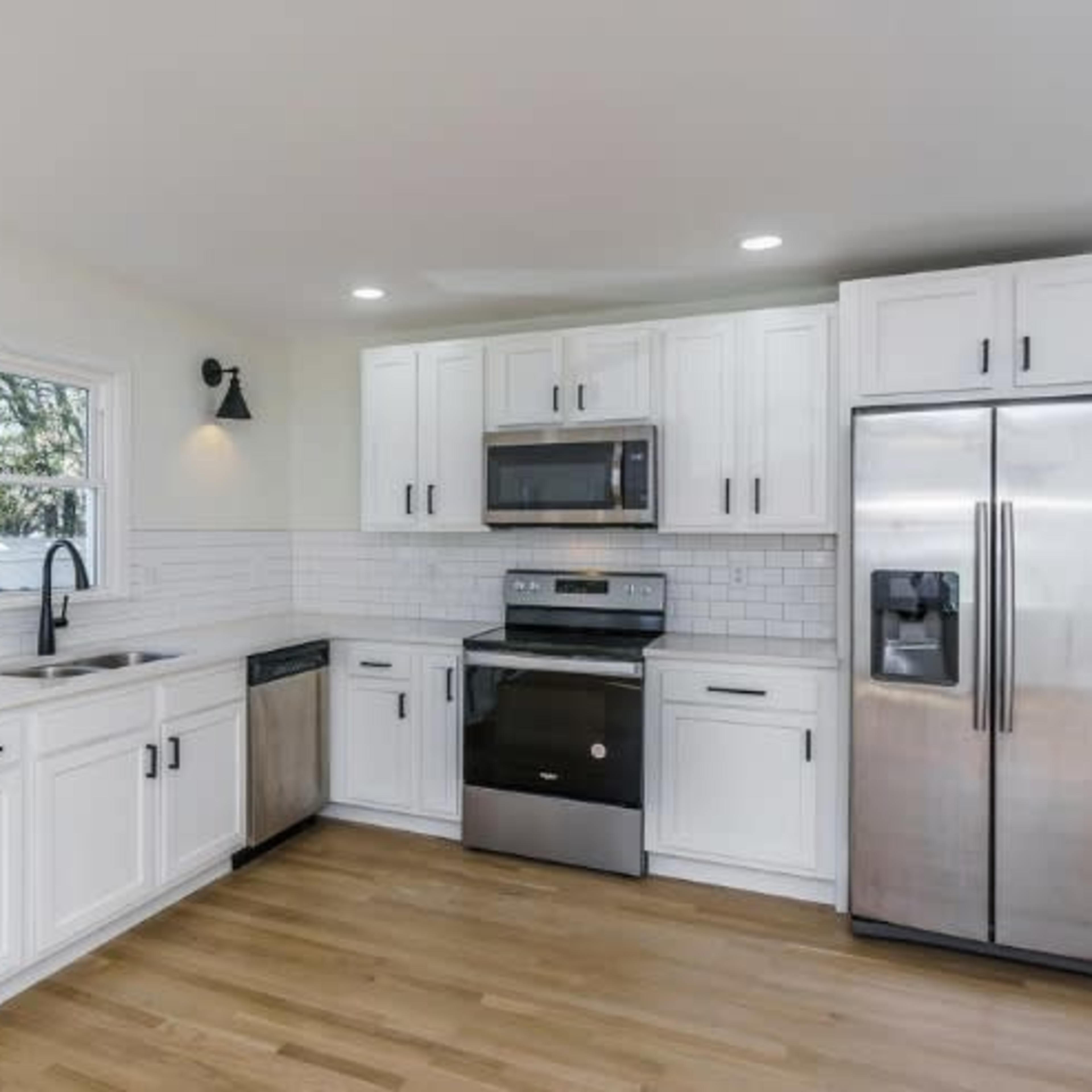 A modern kitchen features white cabinetry, stainless steel appliances, and a wood floor, with a large window providing natural light.