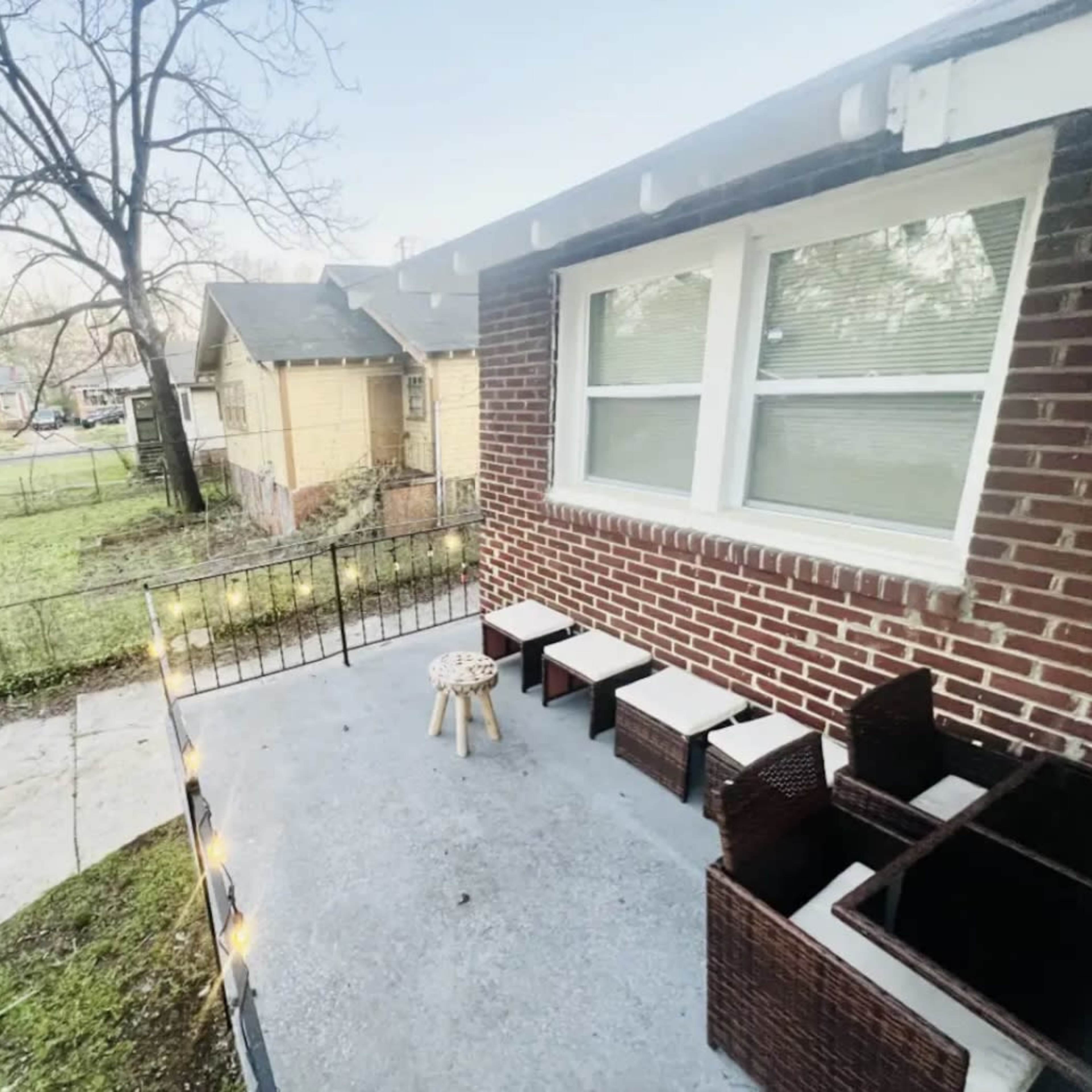 A small patio with a concrete surface, featuring a set of dark furniture and a wooden stool, adjacent to a grassy area and neighboring houses.