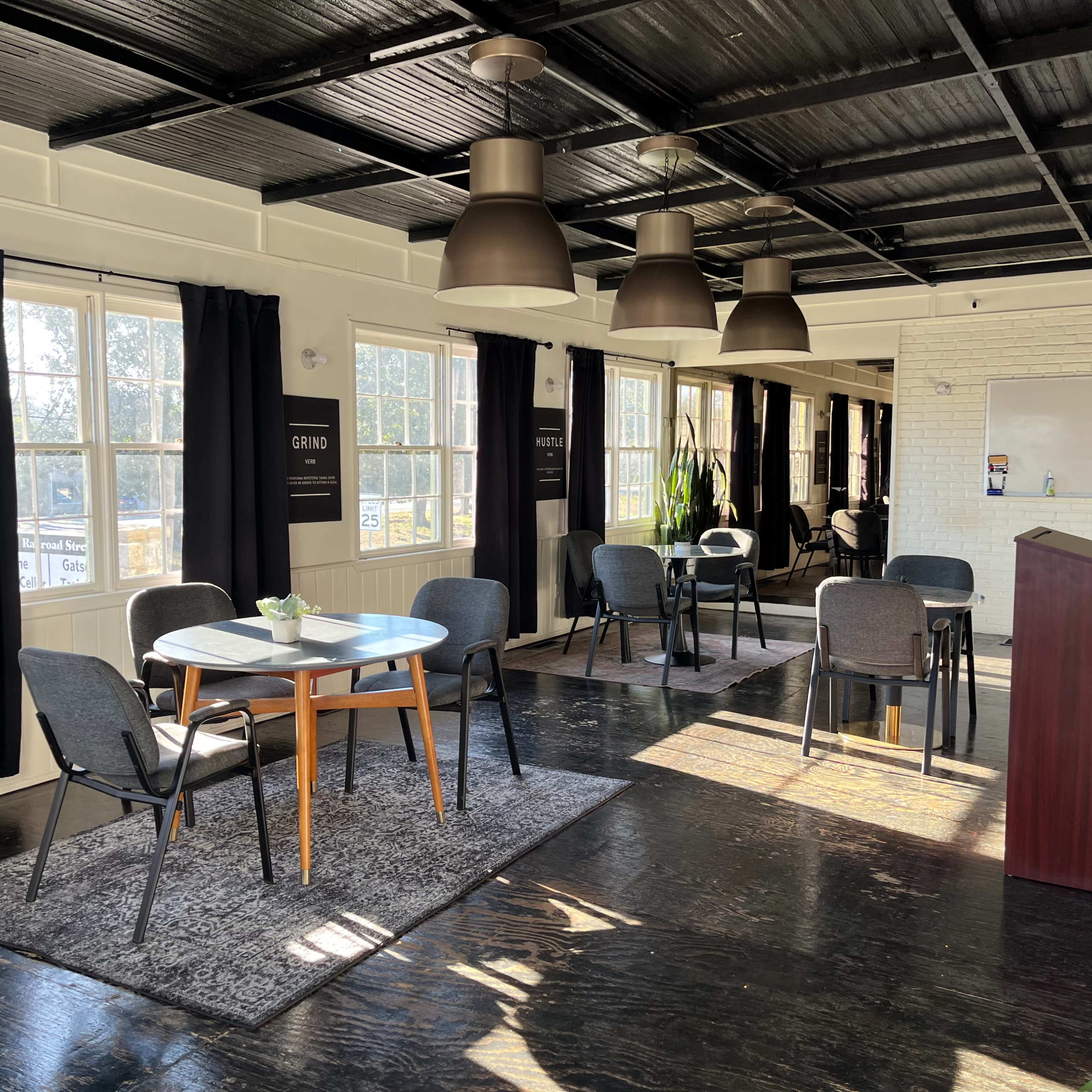 A sunlit room with several tables and chairs arranged around a central area, featuring large windows and a podium at one end.