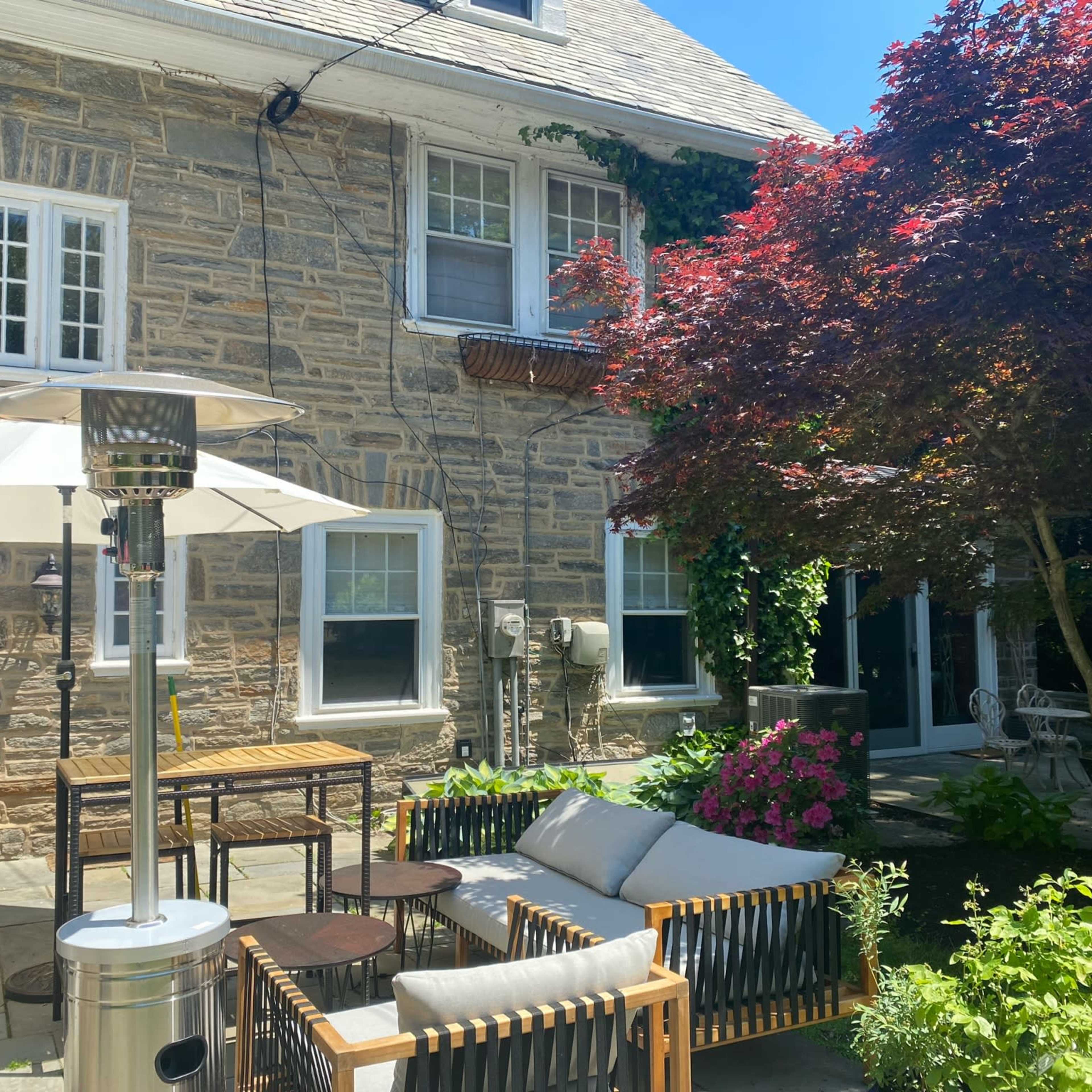 A stone house with green landscaping, featuring a patio area with seating, a heater, and a table beneath an umbrella.