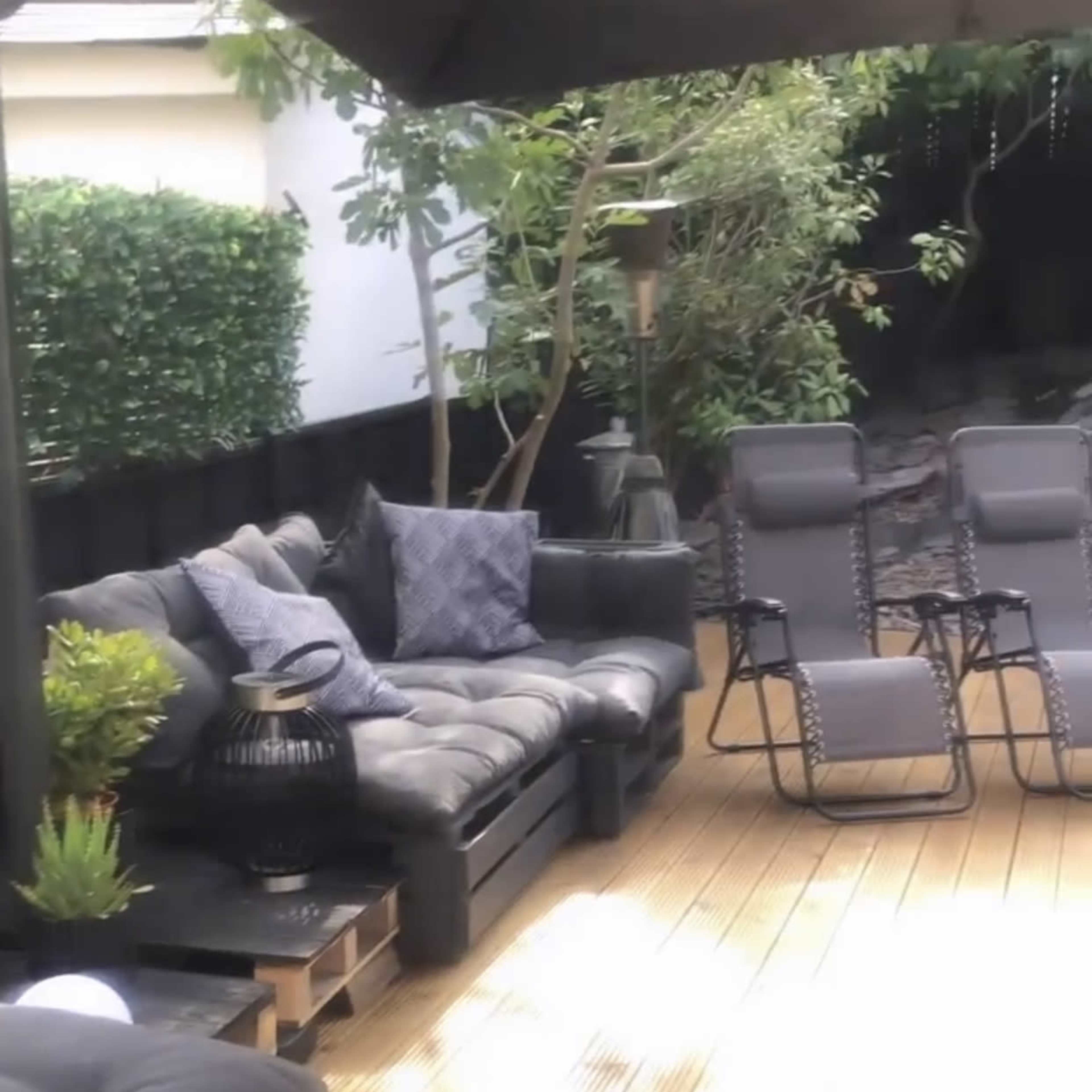 A patio with black outdoor furniture, including a sectional sofa and reclining chairs, set on a wooden deck surrounded by greenery.