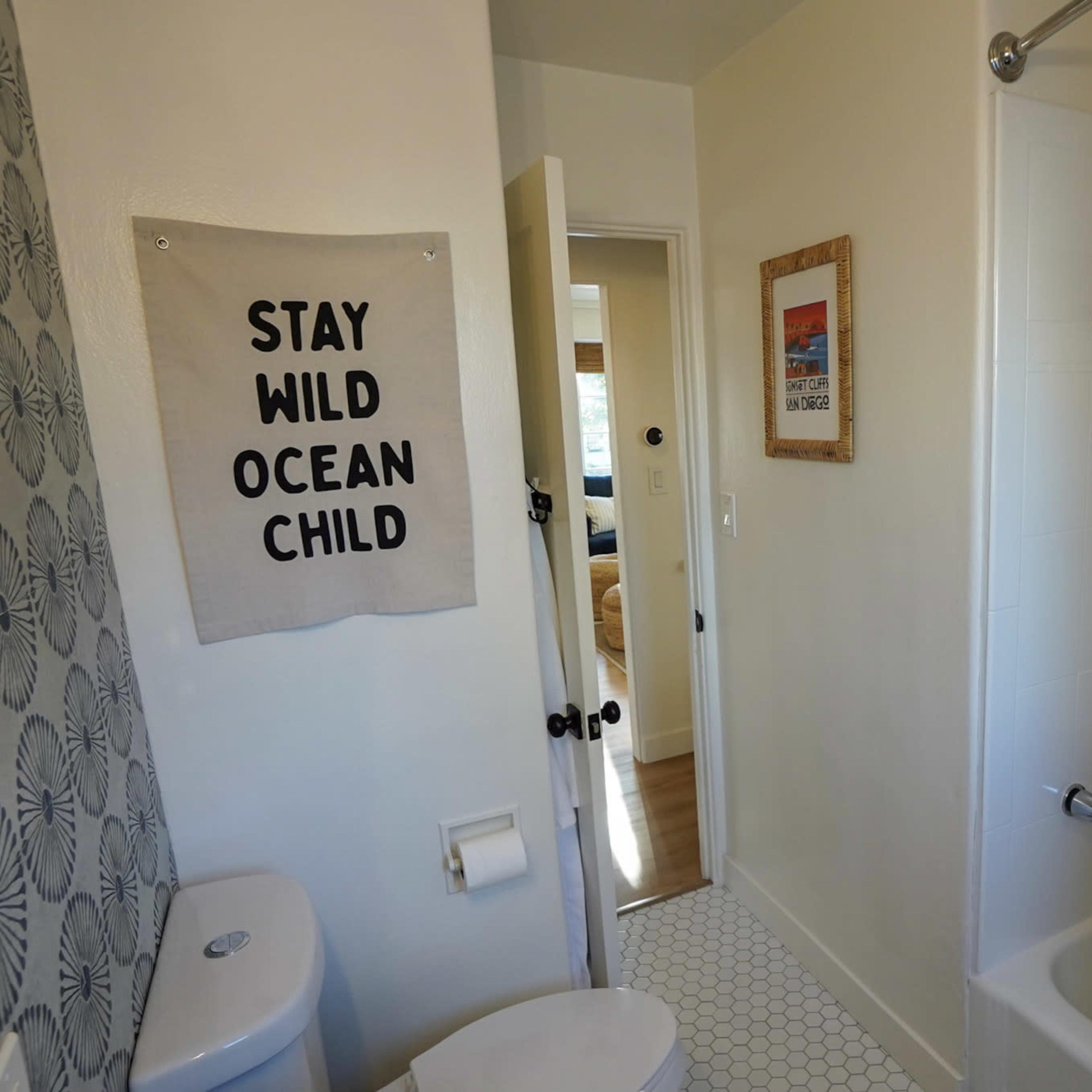 A bathroom with a shower, a toilet, and a wall hanging that reads "STAY WILD OCEAN CHILD."