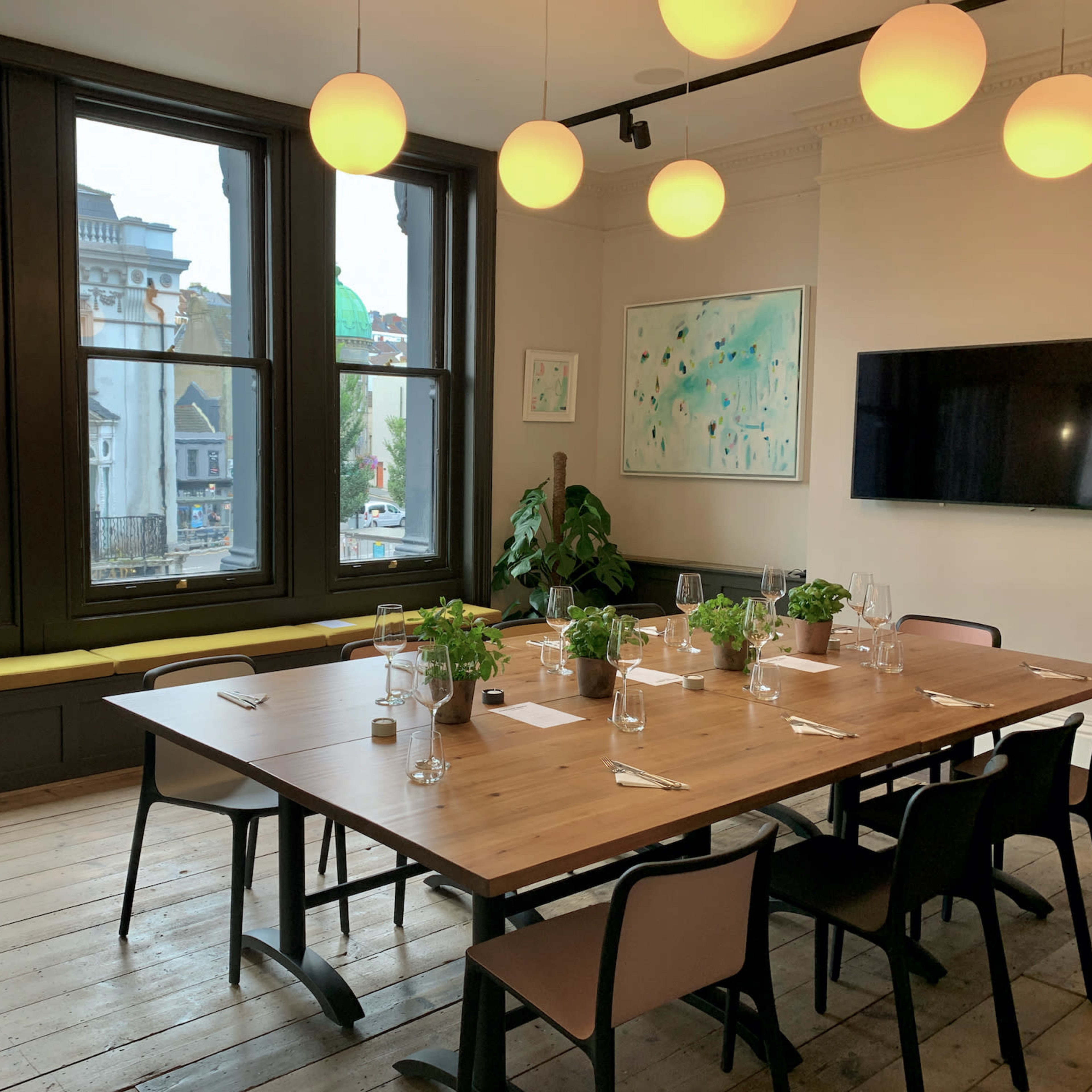 A dining room features a large wooden table set with plates and glasses, surrounded by black chairs, with large windows offering a view of the street outside.