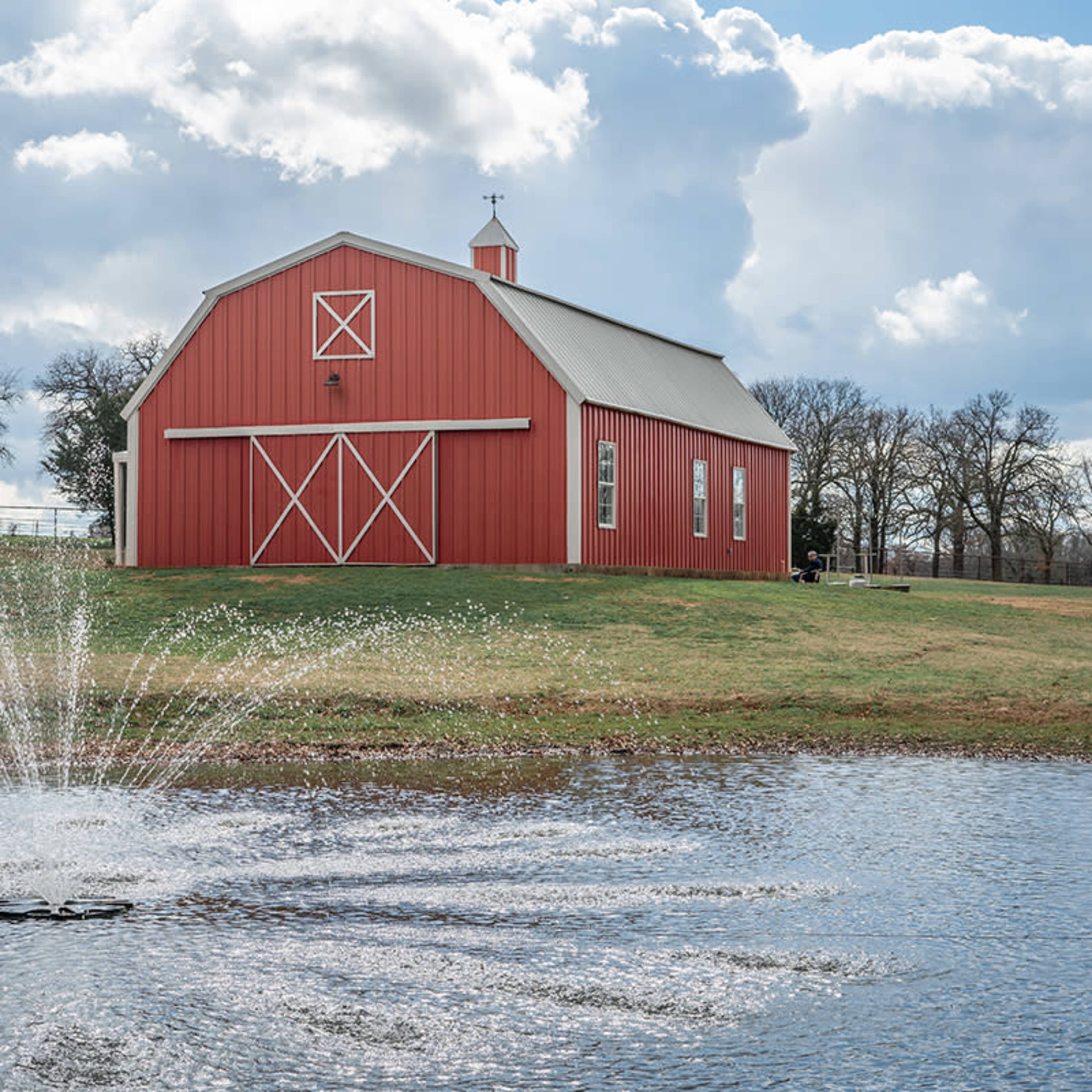A red barn stands beside a pond with a fountain, surrounded by trees and a cloudy sky.