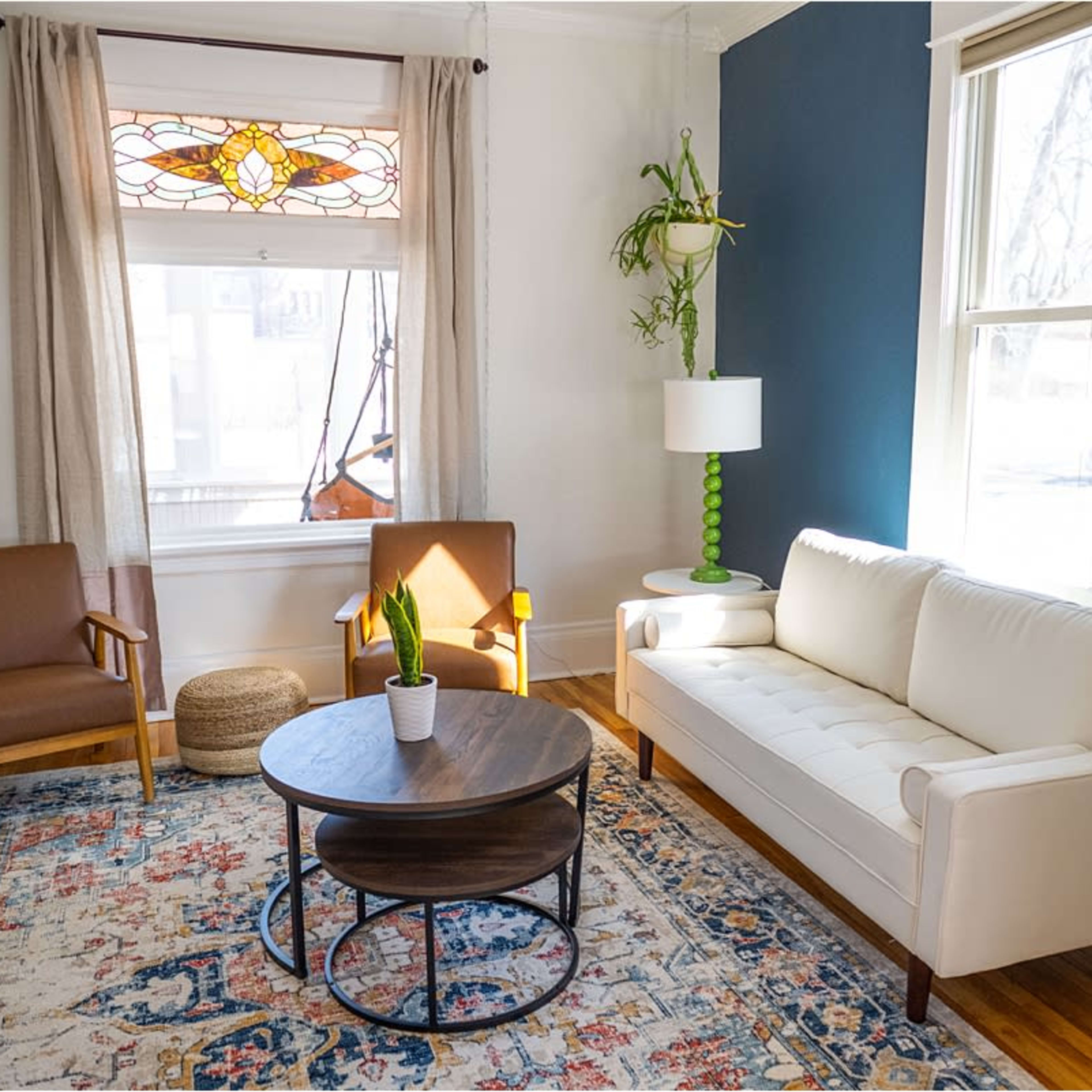 The living room features a white sofa, two brown armchairs, a round coffee table, and large windows with sheer curtains.