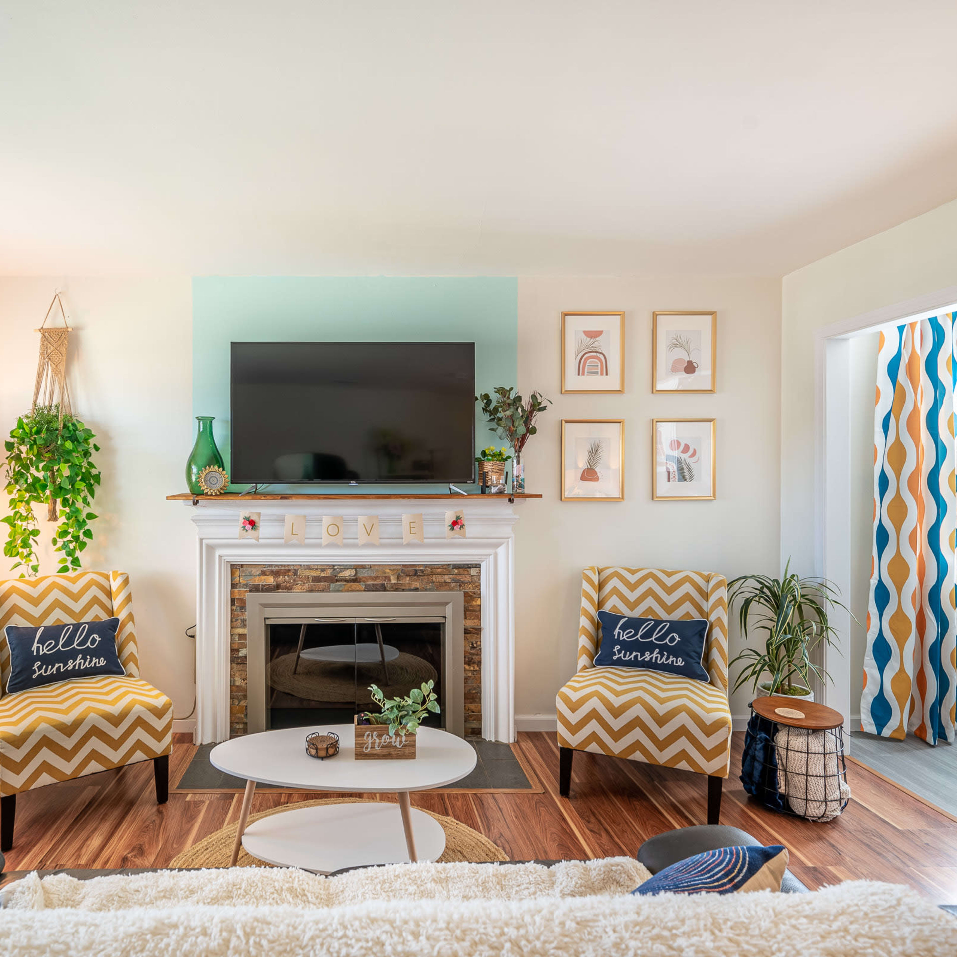 A bright living room featuring a fireplace with a mounted television above, flanked by two patterned chairs and decorative plants.