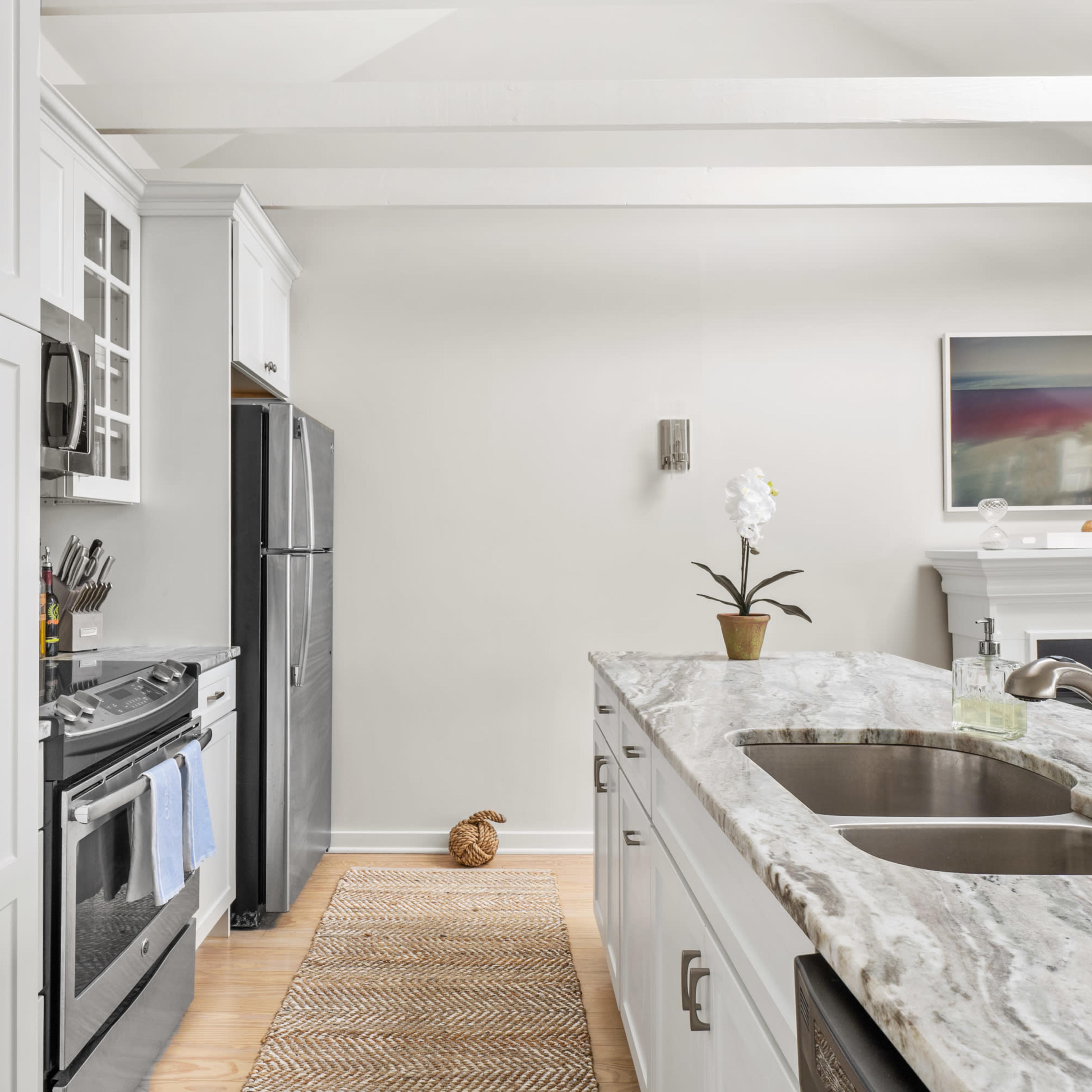A modern kitchen features white cabinetry, a gray marble countertop, and a small potted plant on the counter, with a view of an adjacent living area and a fireplace.