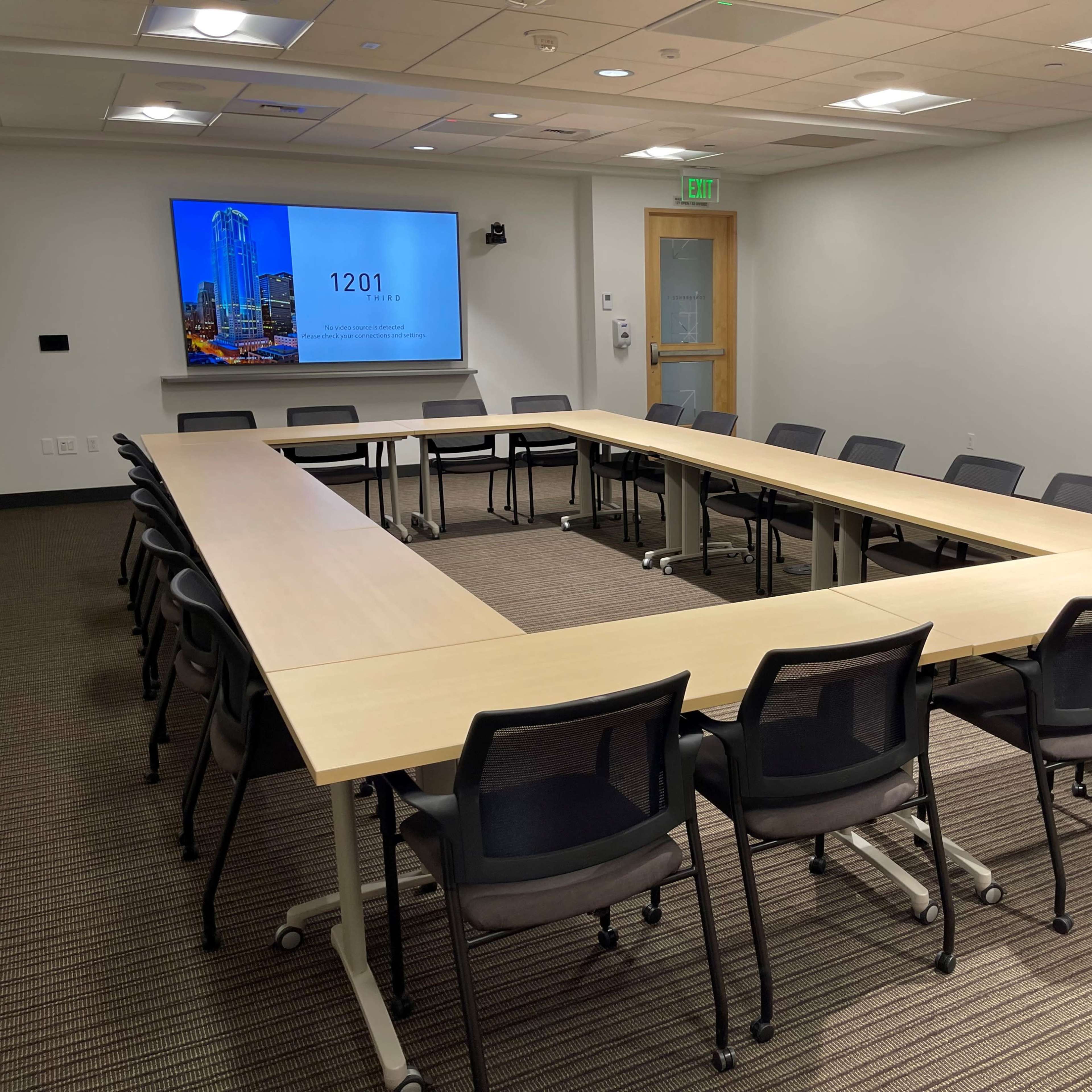 A modern conference room features a long, rectangular table surrounded by black chairs, with a presentation screen displaying "1201" on one wall.