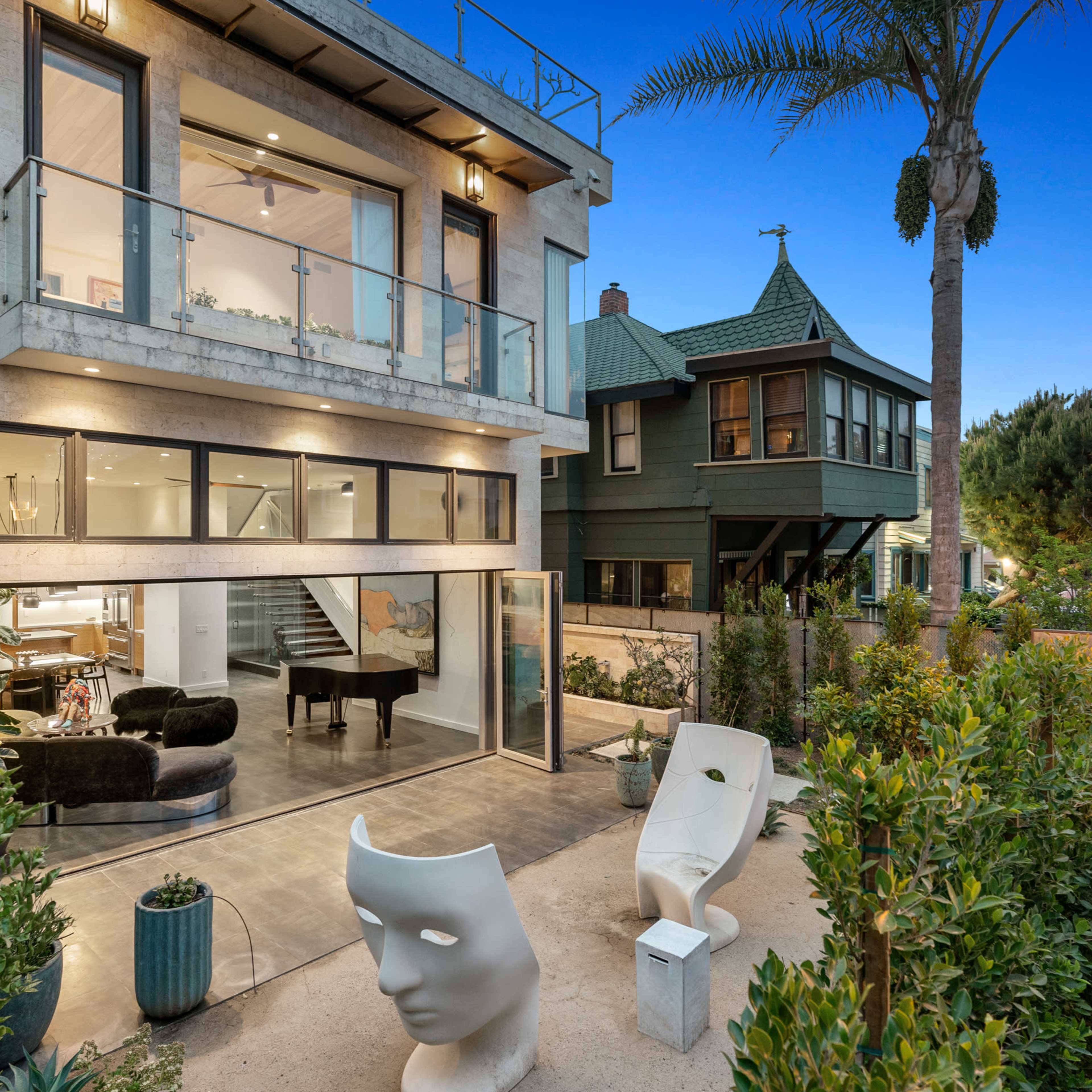 A modern two-story house with large windows and a balcony, surrounded by landscaped greenery and featuring a unique sculpture in the foreground.