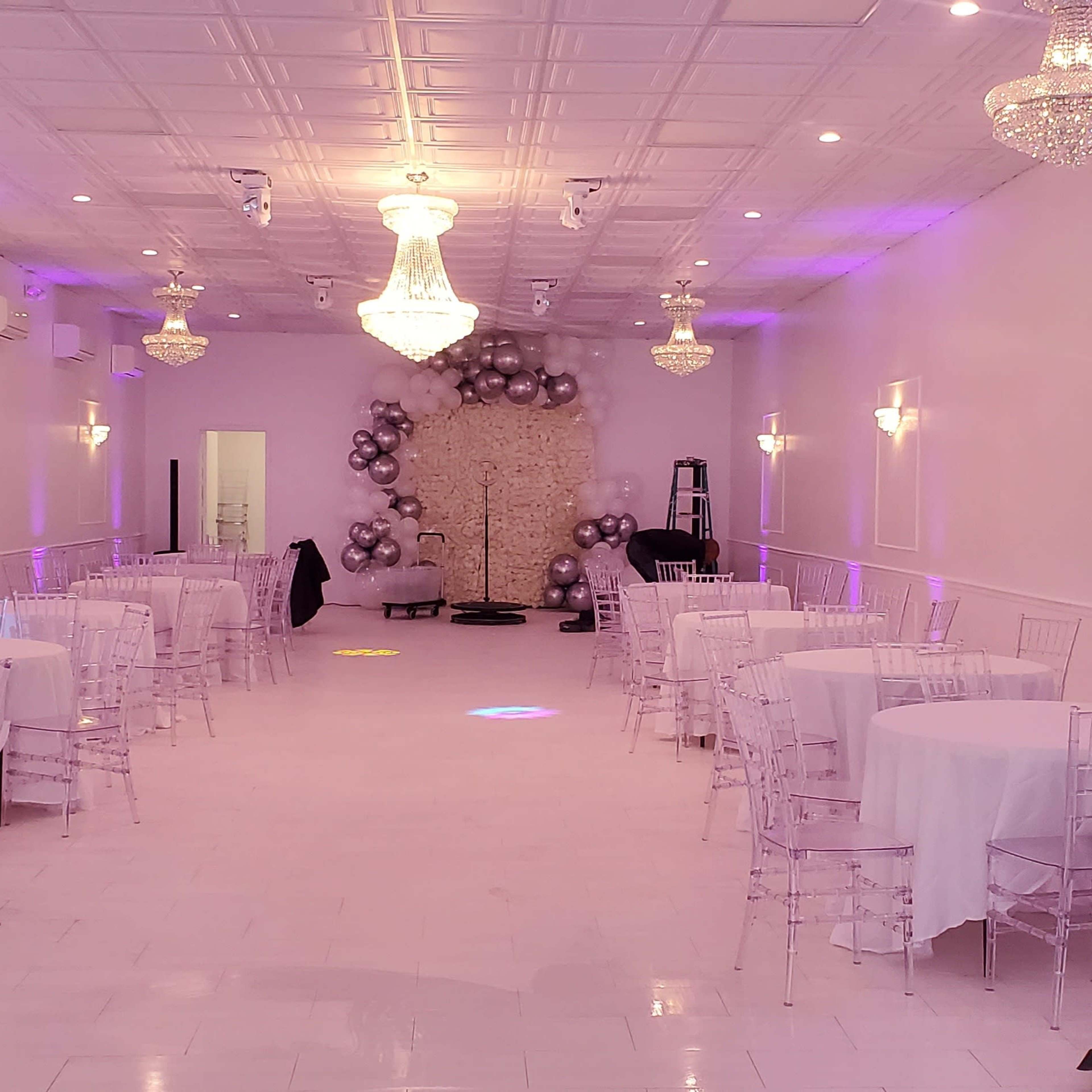 A spacious event hall with white walls, round tables covered in white cloth, and elegant chandeliers, illuminated by soft purple lighting.