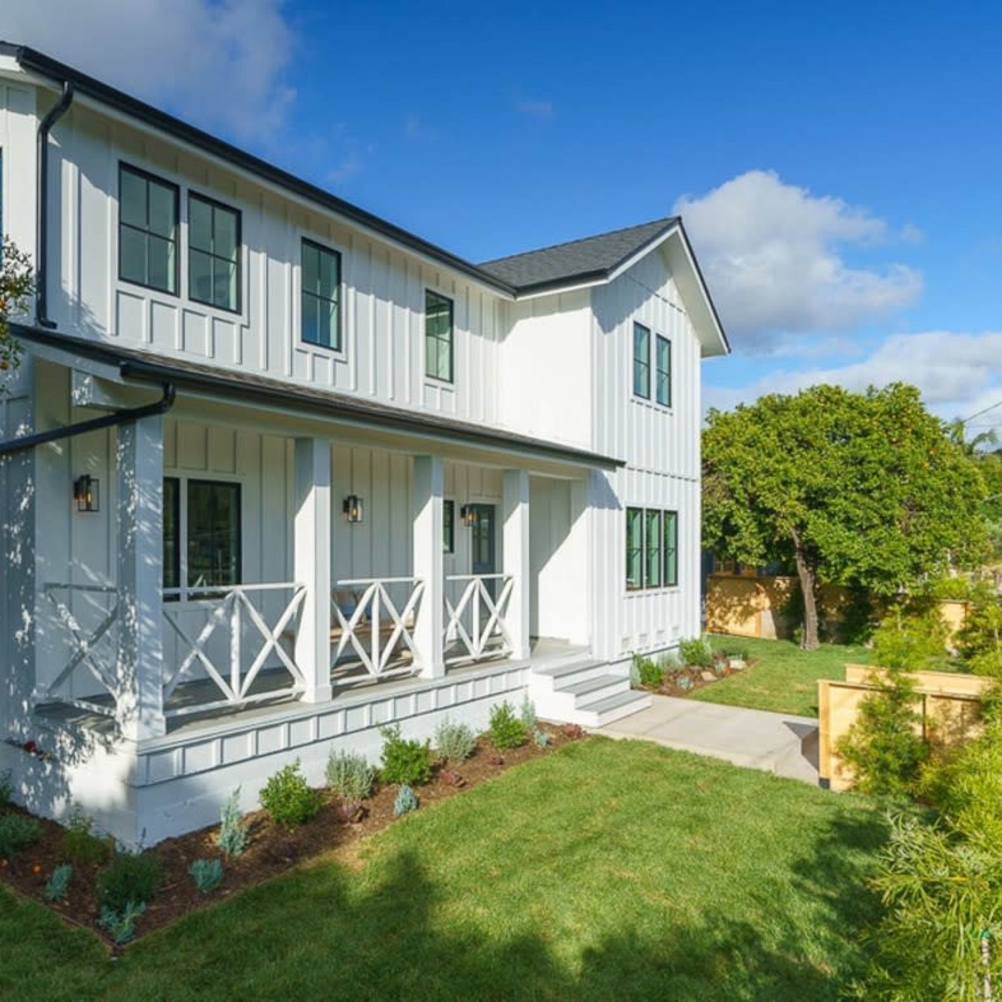 A modern two-story white wooden house with a porch and a landscaped yard featuring a citrus tree.