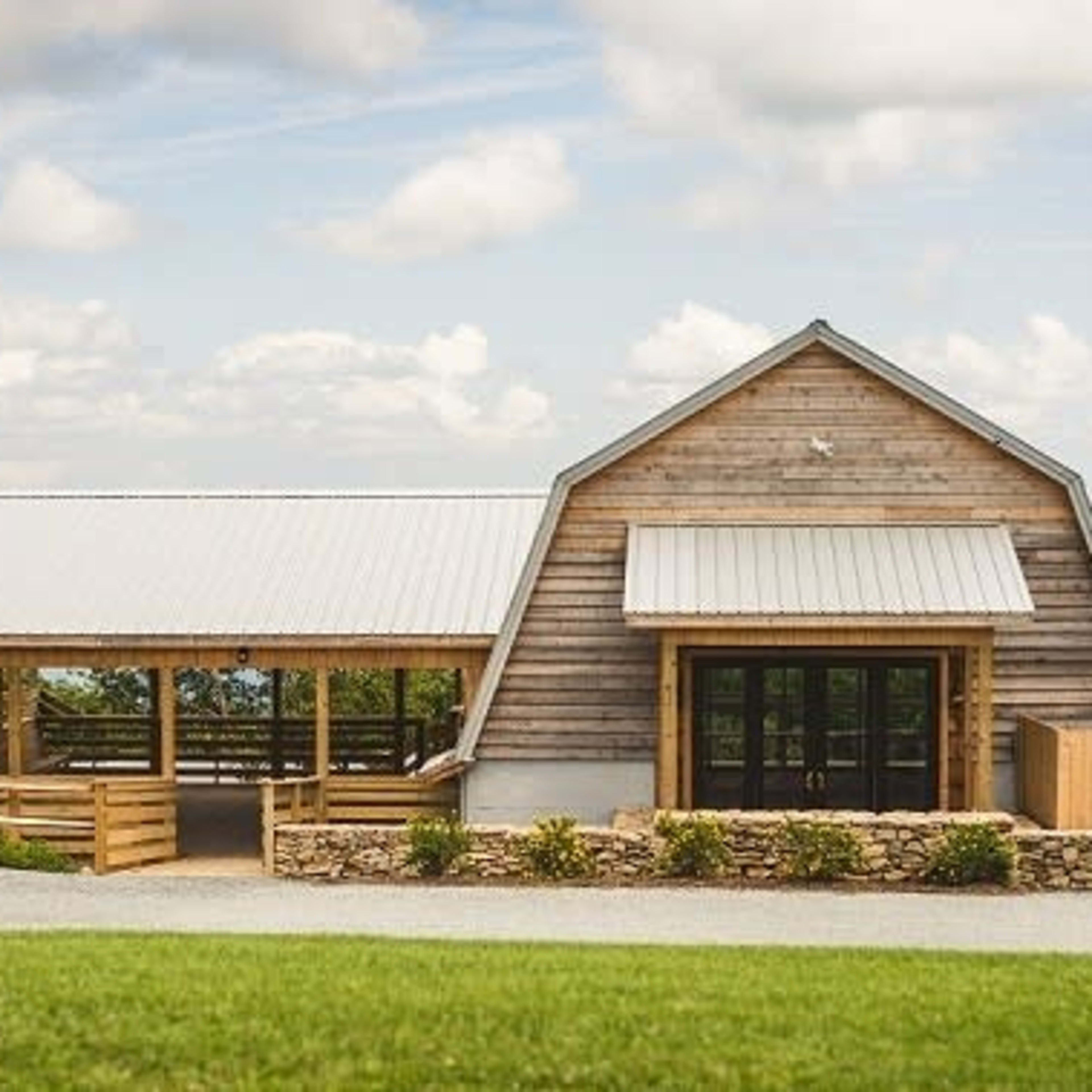 A wooden structure with a gabled roof, featuring large glass doors and a covered porch, situated on a grassy area with mountains in the background.