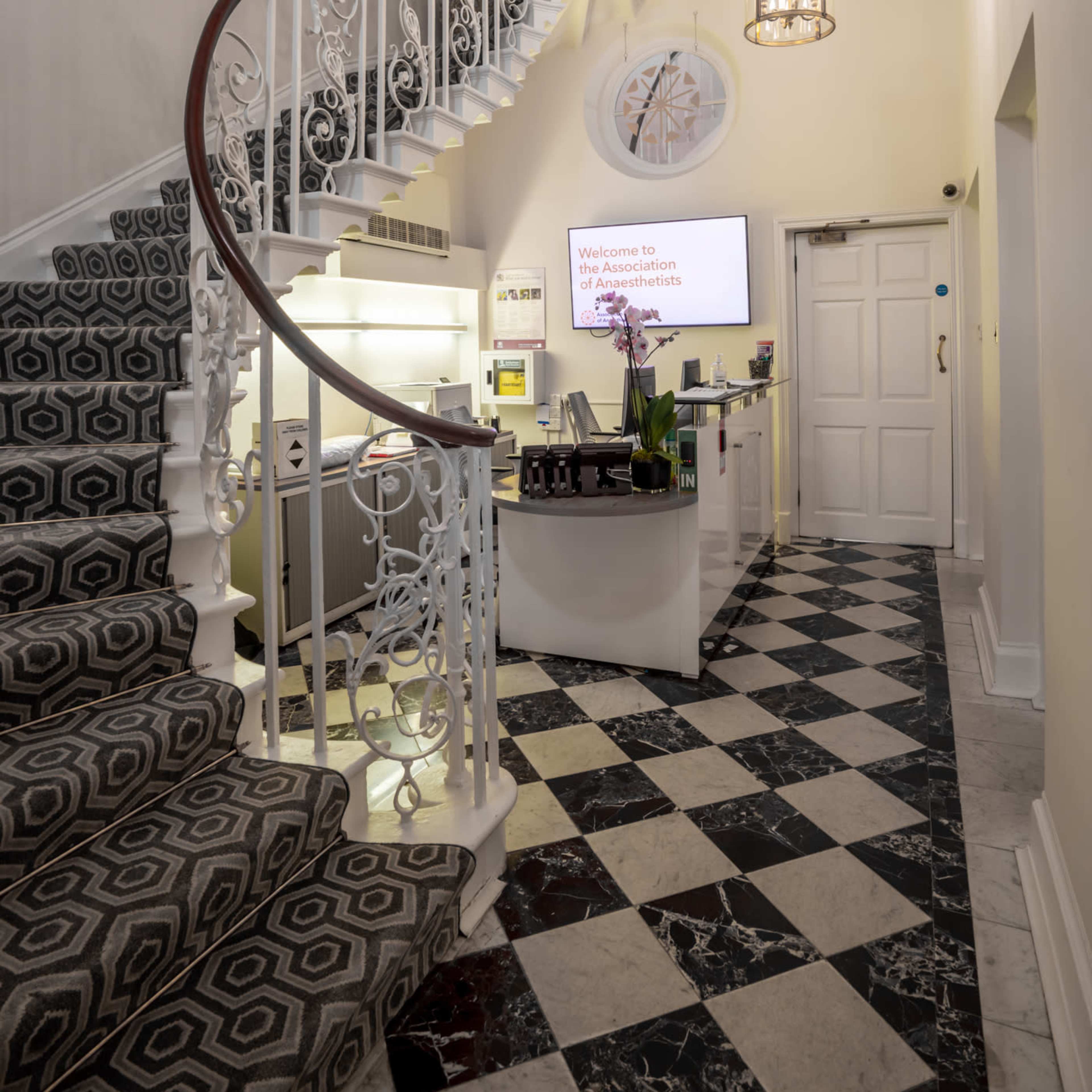 A well-lit reception area featuring a curved staircase with decorative railings, a modern front desk, and a patterned tiled floor.