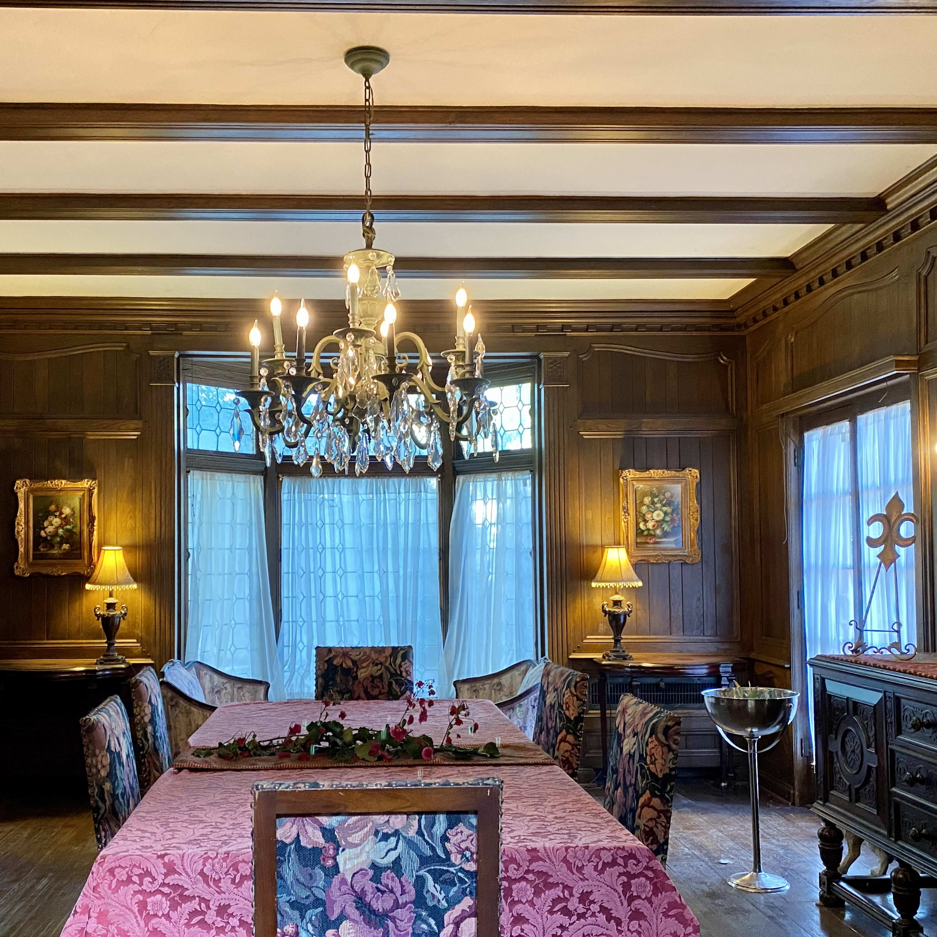 An elegantly furnished dining room featuring a long table with a pink tablecloth, surrounded by upholstered chairs, and illuminated by a crystal chandelier.