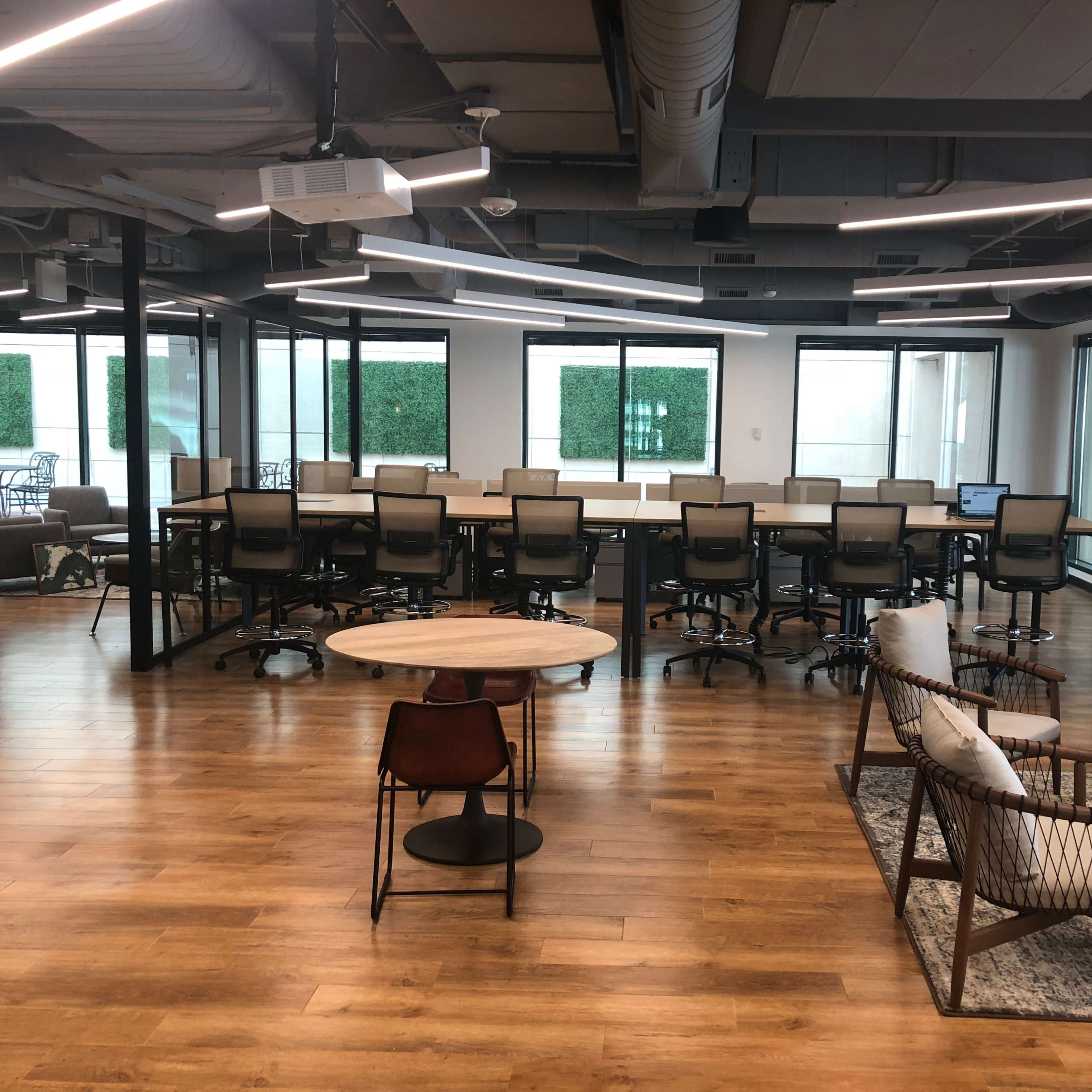 A modern office space with a large table surrounded by chairs, a lounge area with seating, and multiple large windows.