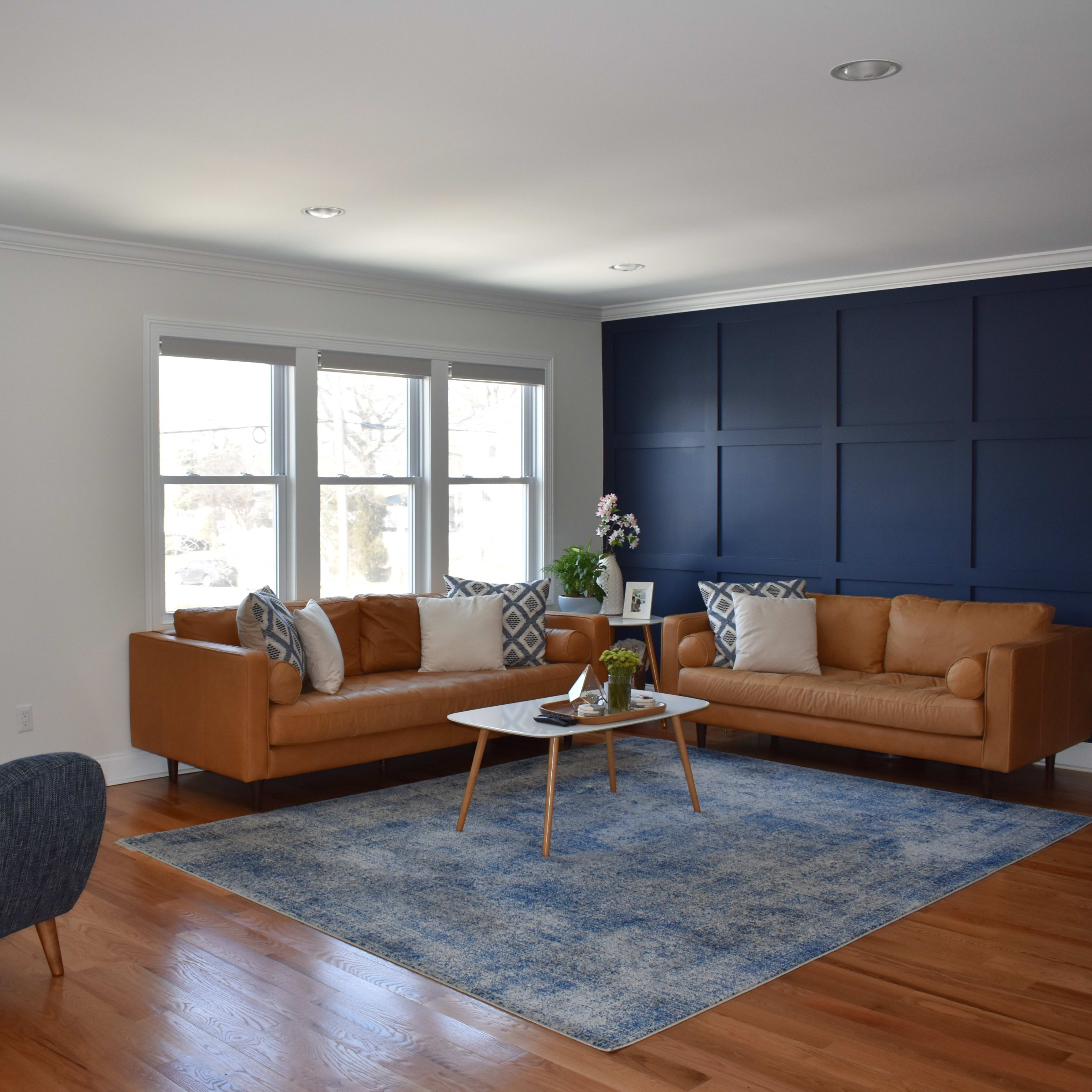 A contemporary living room with a brown leather sofa, a blue area rug, and a dark blue accent wall, complemented by a coffee table and two upholstered chairs.