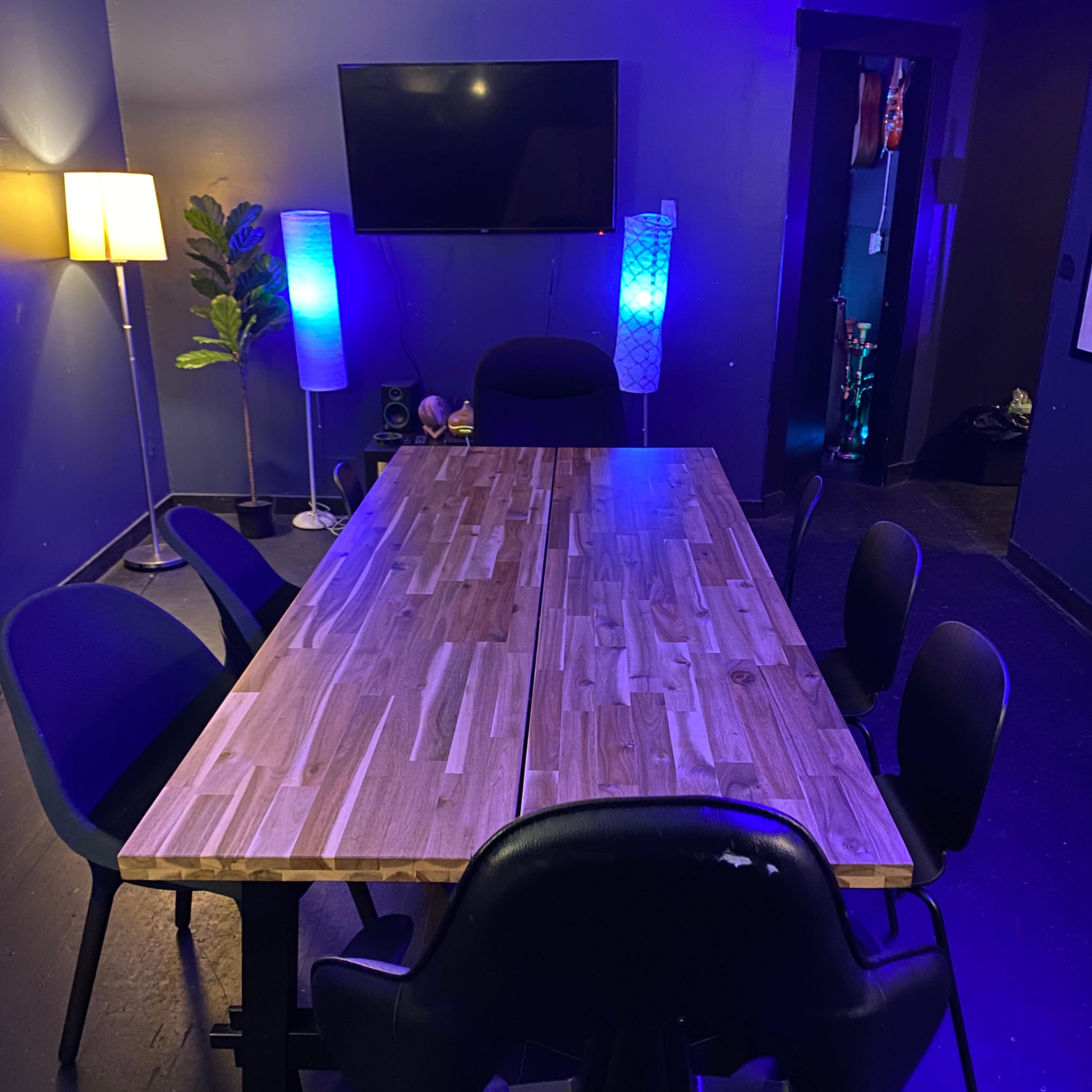 A long wooden table surrounded by black chairs in a dimly lit room with blue lighting and a television on the wall.