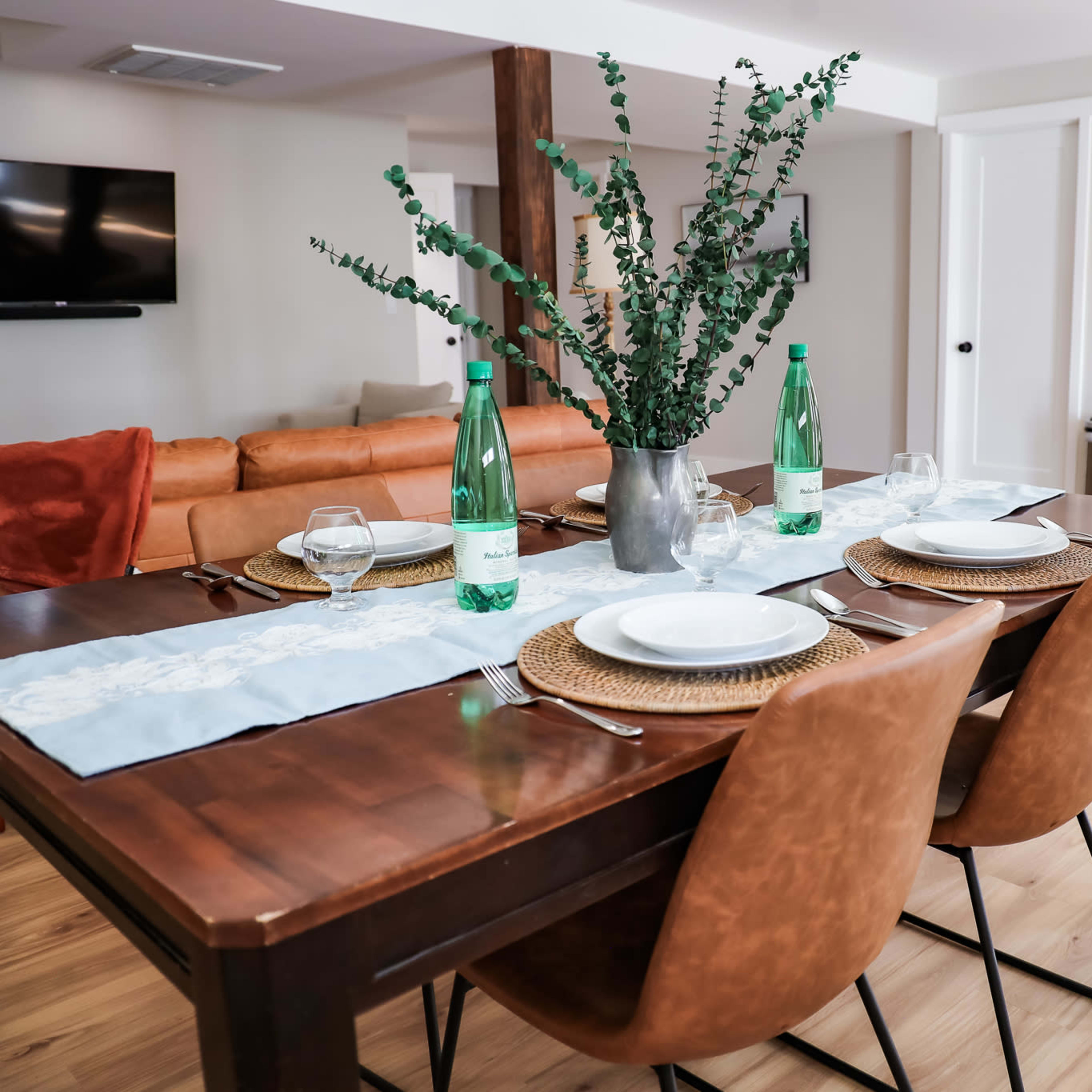 A dining table is set for a meal with glassware and plates, accompanied by a vase of greenery, in a bright and open kitchen-living space featuring a couch and a television.