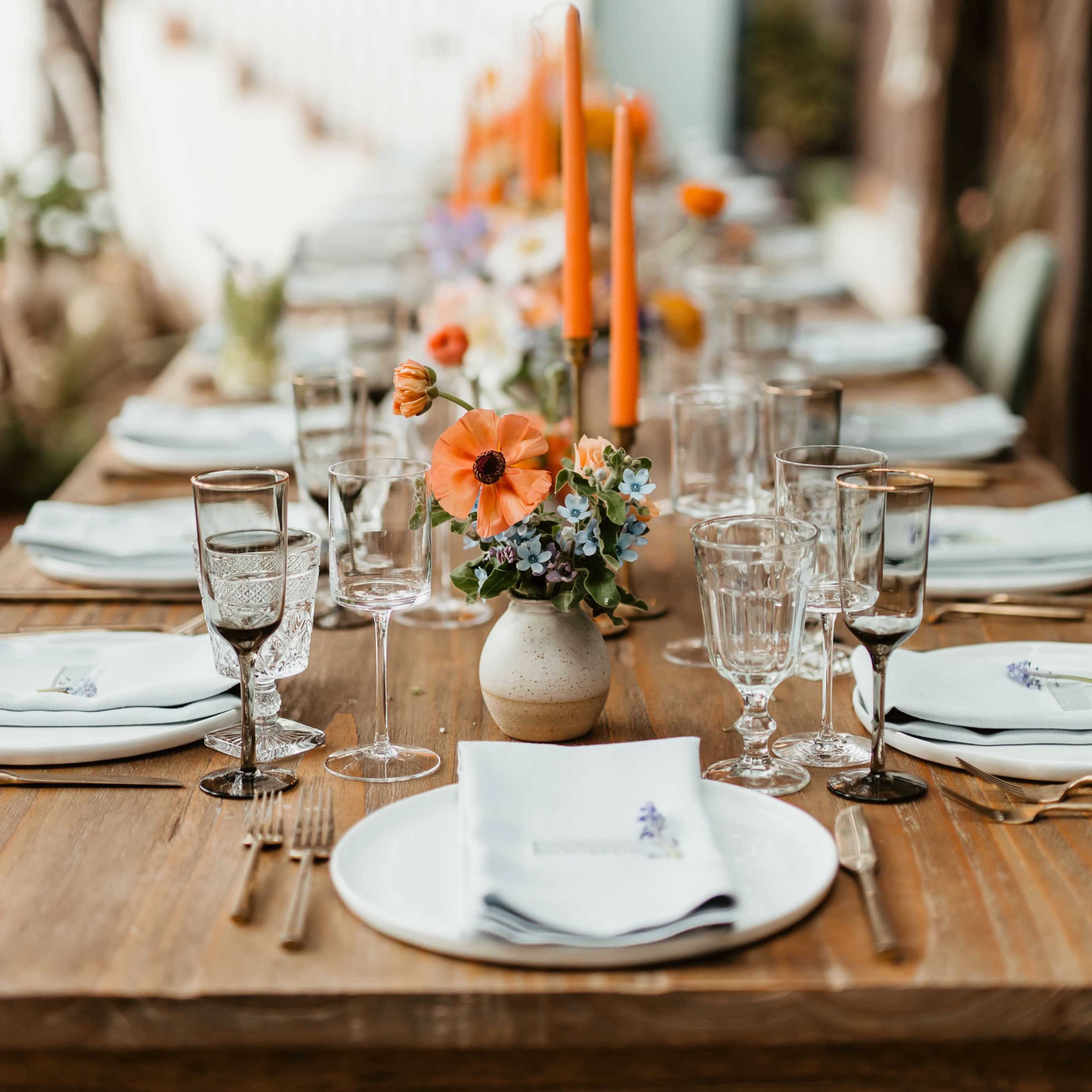 A long wooden table is set for a meal under a canopy of green foliage and blooming flowers.