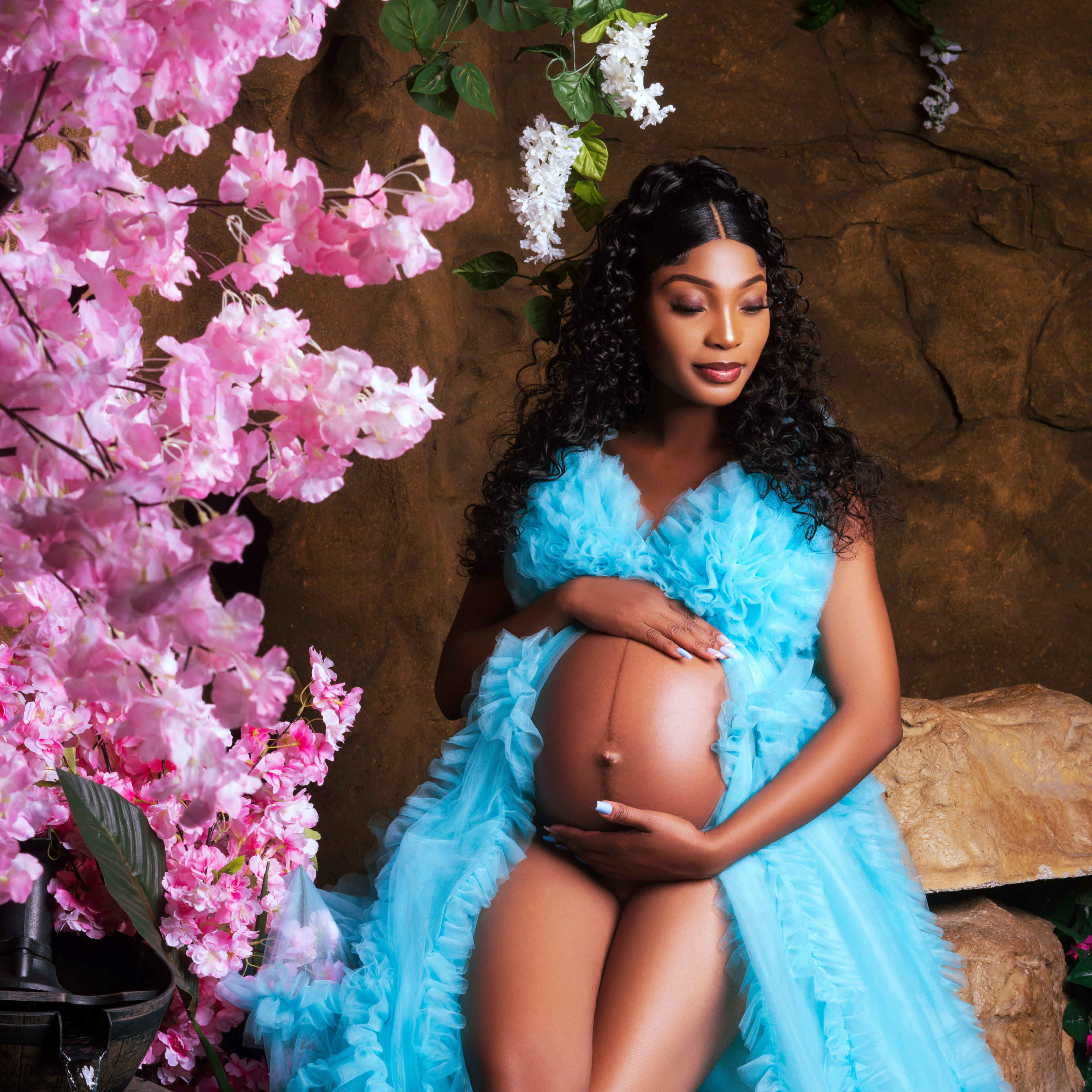 A woman in a blue dress gently holds her pregnant belly while seated among pink flowers and decorative rocks.