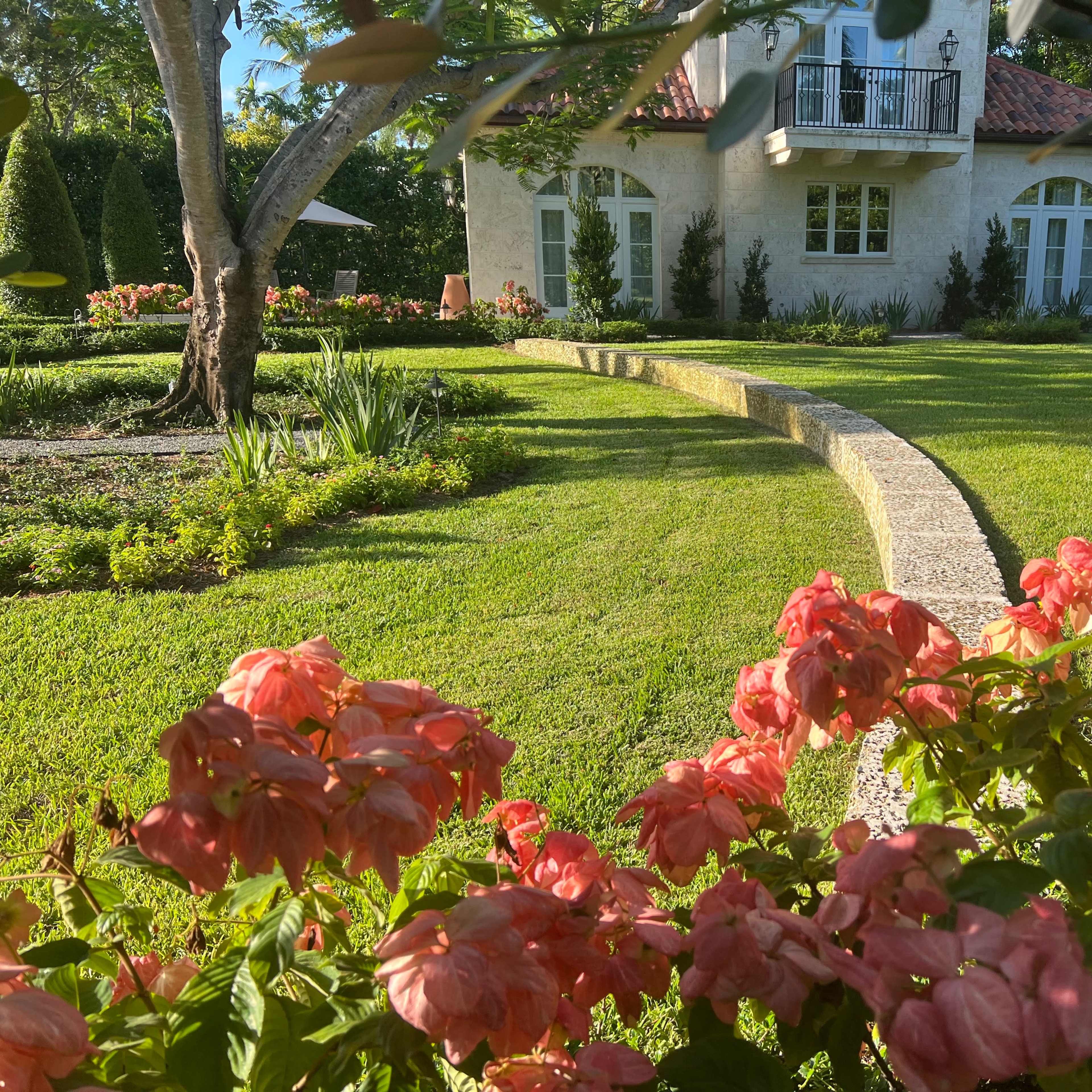 A curved stone path leads through a manicured lawn to a house surrounded by landscaped gardens and blooming pink flowers.