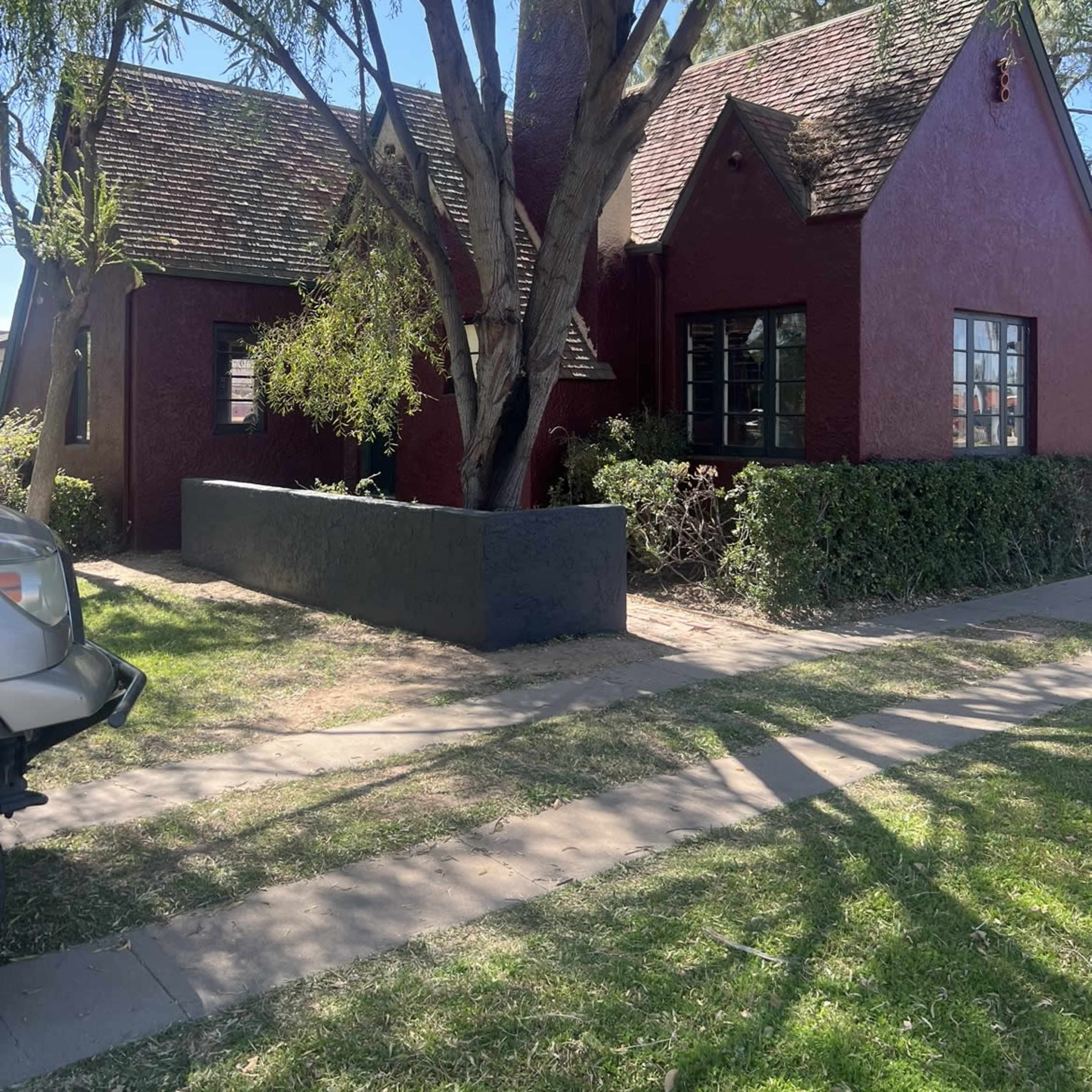 A red cottage with a steep roof and surrounded by greenery stands on a tree-lined street.