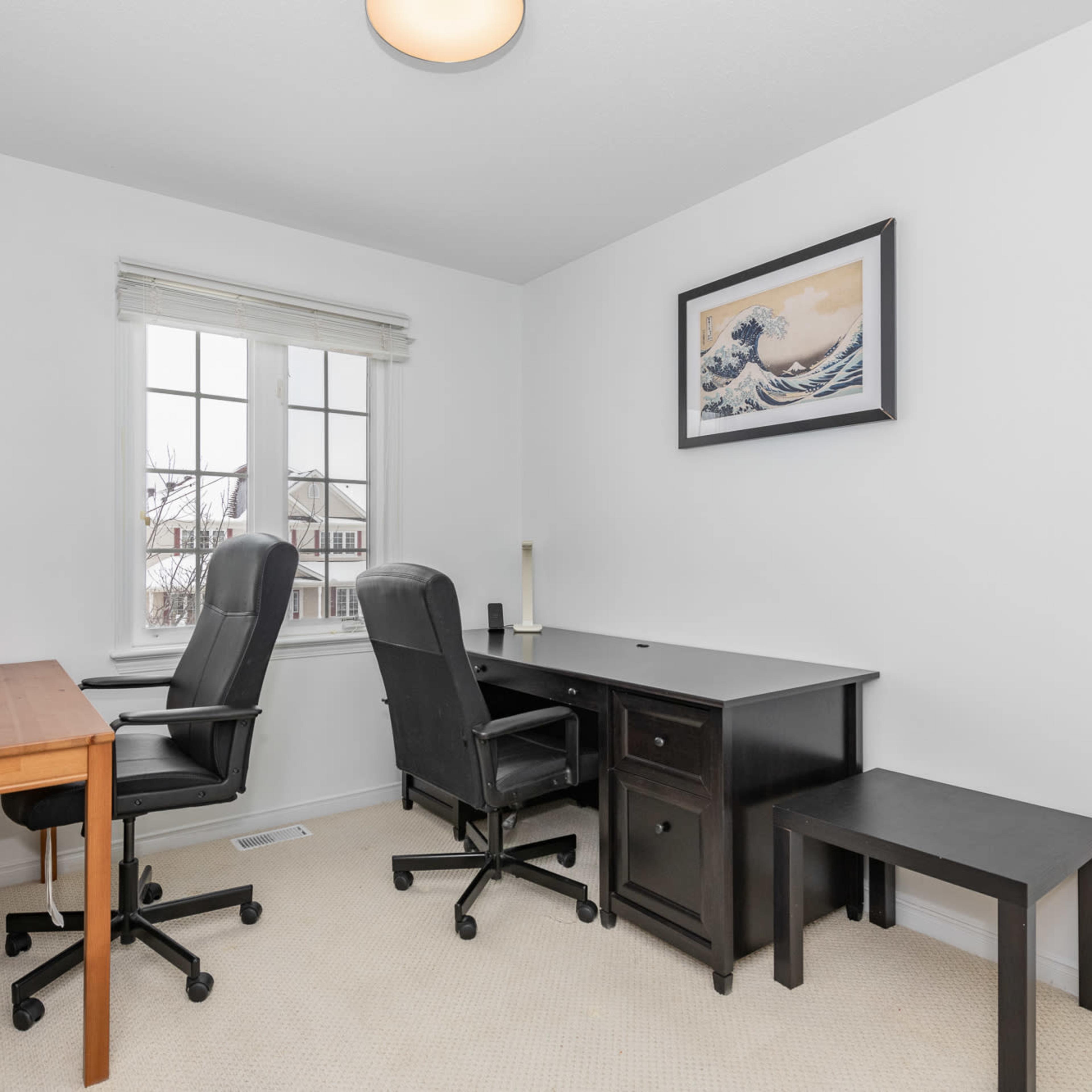 A bright home office with two black chairs, a wooden desk, a black desk with a drawer, and a shelf against the wall.
