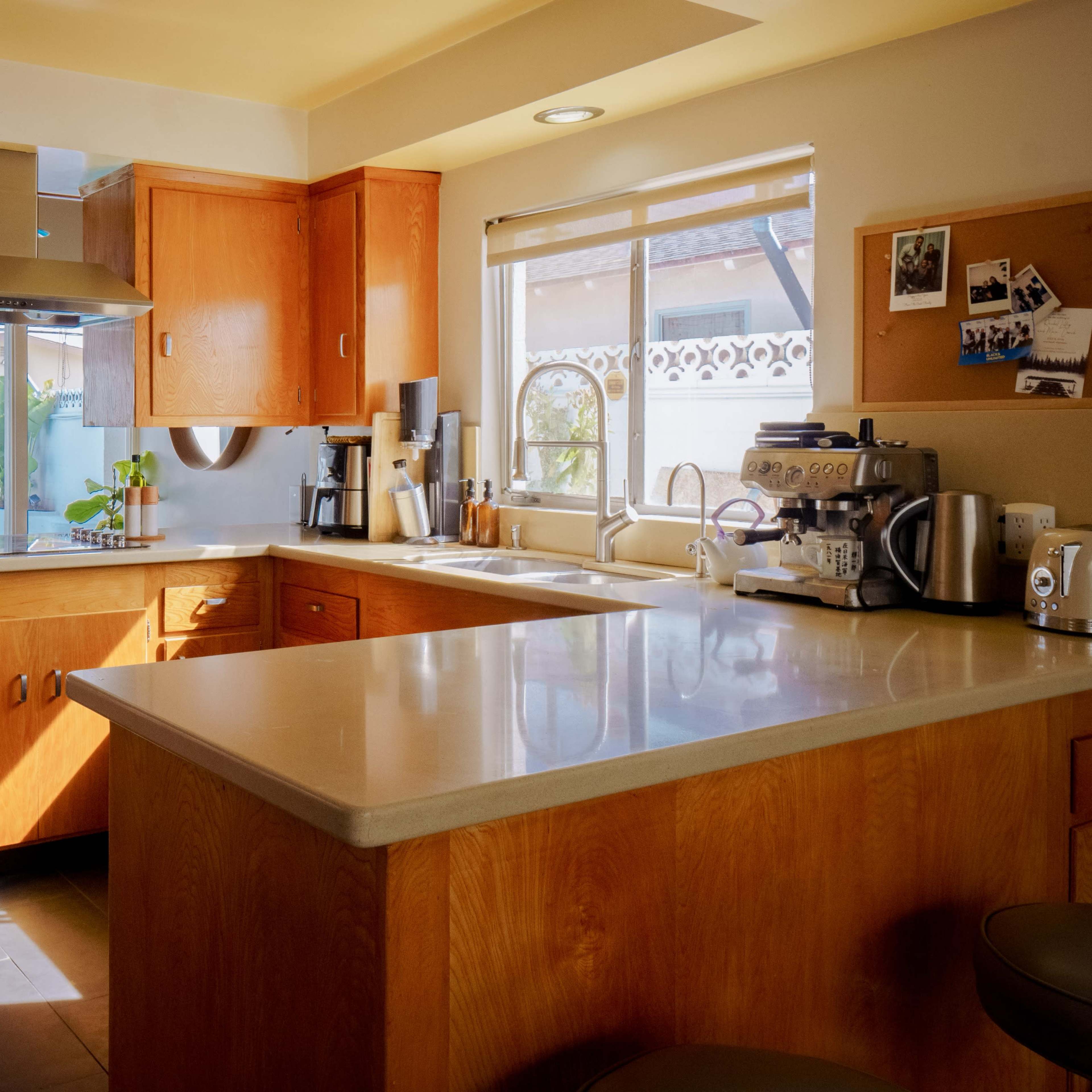 A modern kitchen with wooden cabinets, a stainless steel range hood, and a countertop that features a coffee machine and various kitchen tools, illuminated by natural light.
