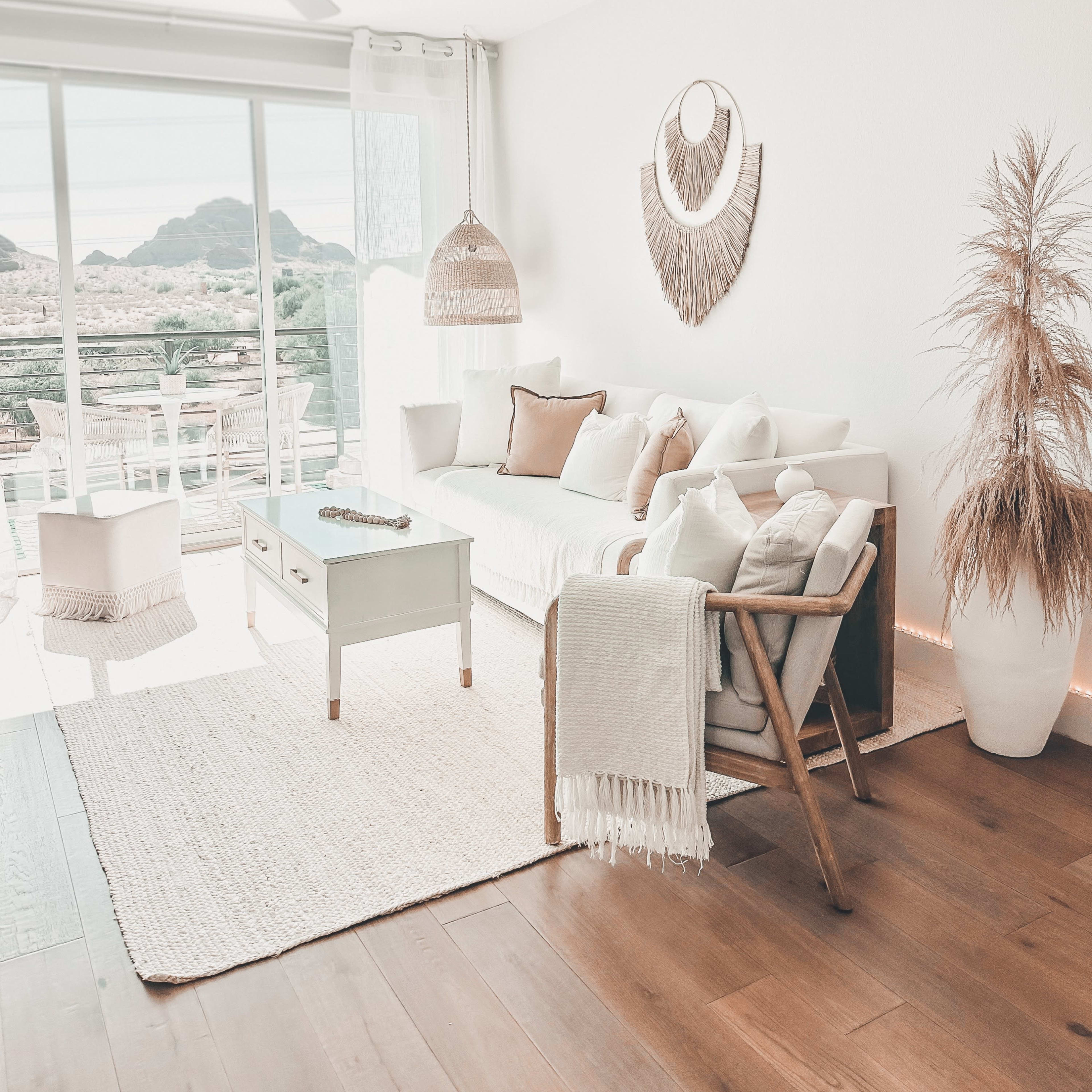 The living room features a light-colored sofa with various cushions, a green coffee table, a woven pendant lamp, and decorative plants, all arranged around a spacious window offering a view of mountains.