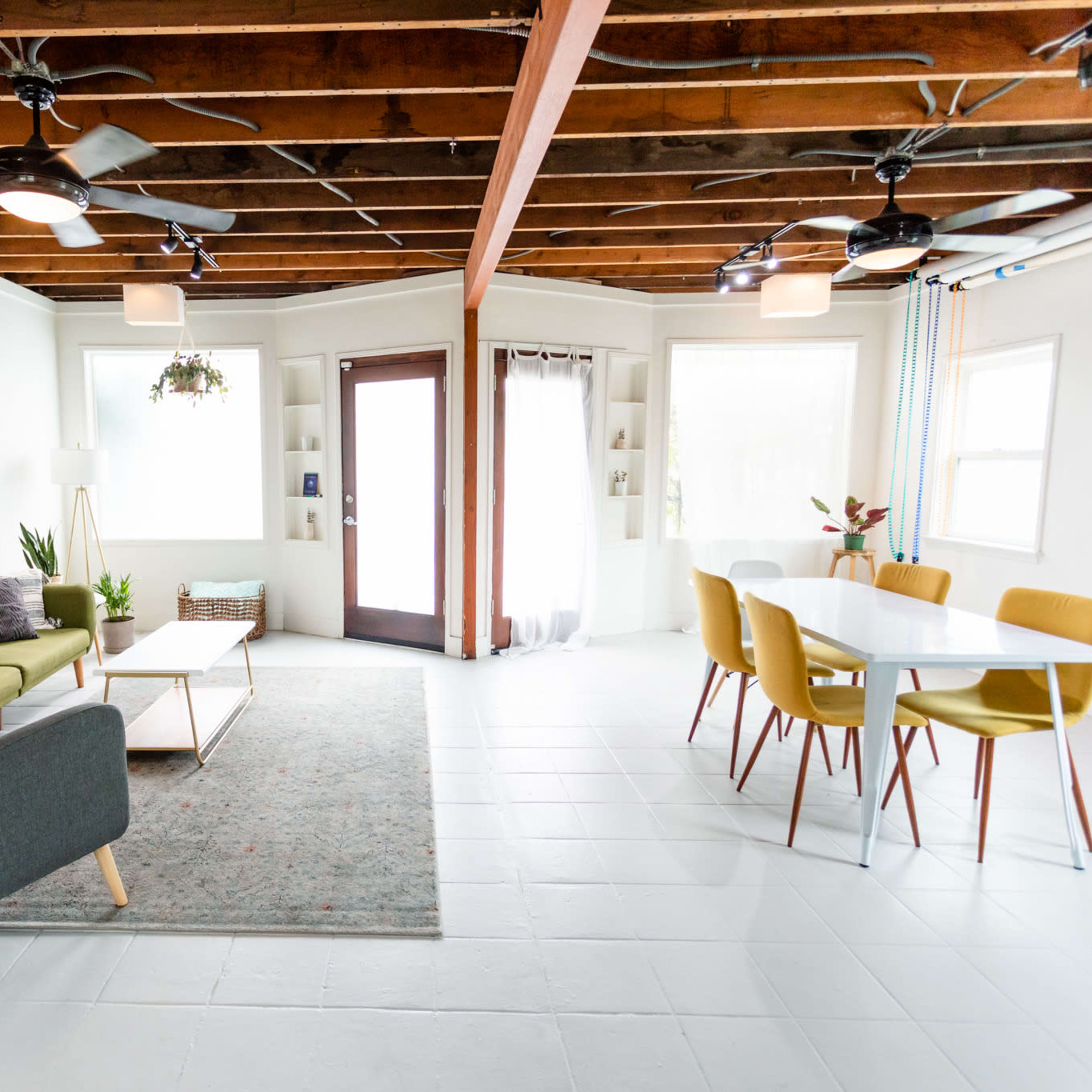 A bright living room with a green couch, a gray armchair, a dining table surrounded by colorful chairs, and large windows letting in natural light.