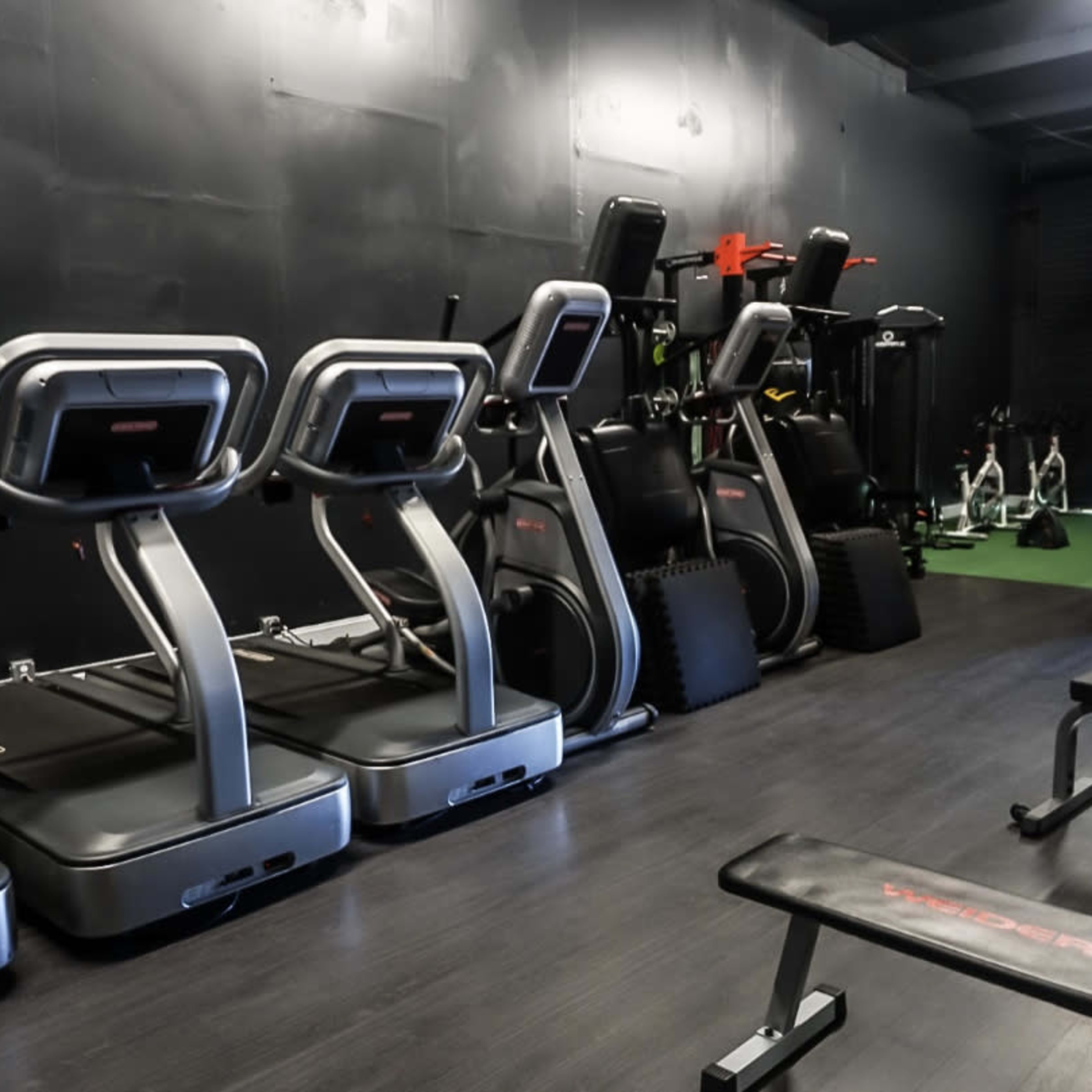 A gym interior featuring multiple treadmills lined up against a black wall, with various exercise equipment stored in the background.