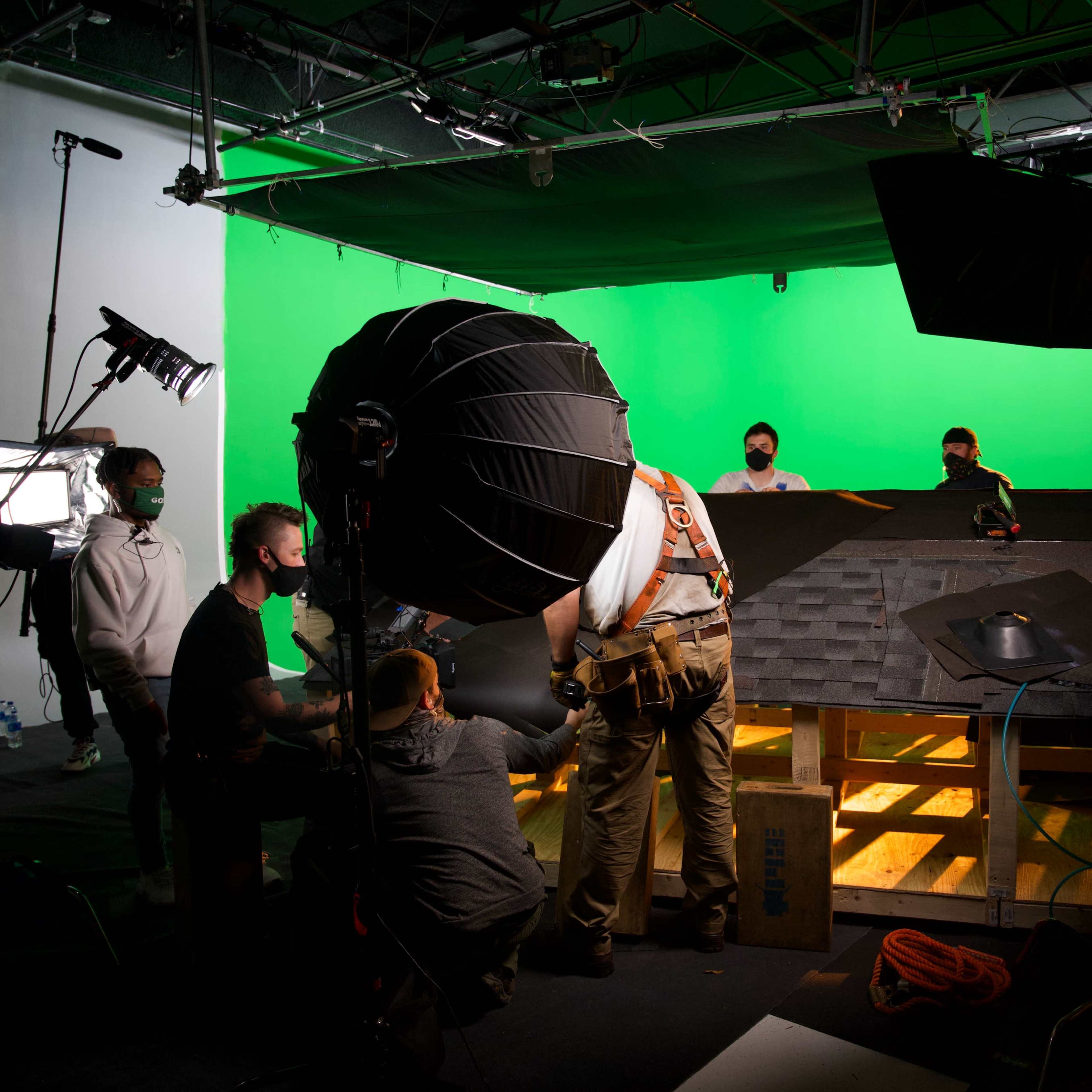A film crew works on a set with a green screen backdrop and lighting equipment.