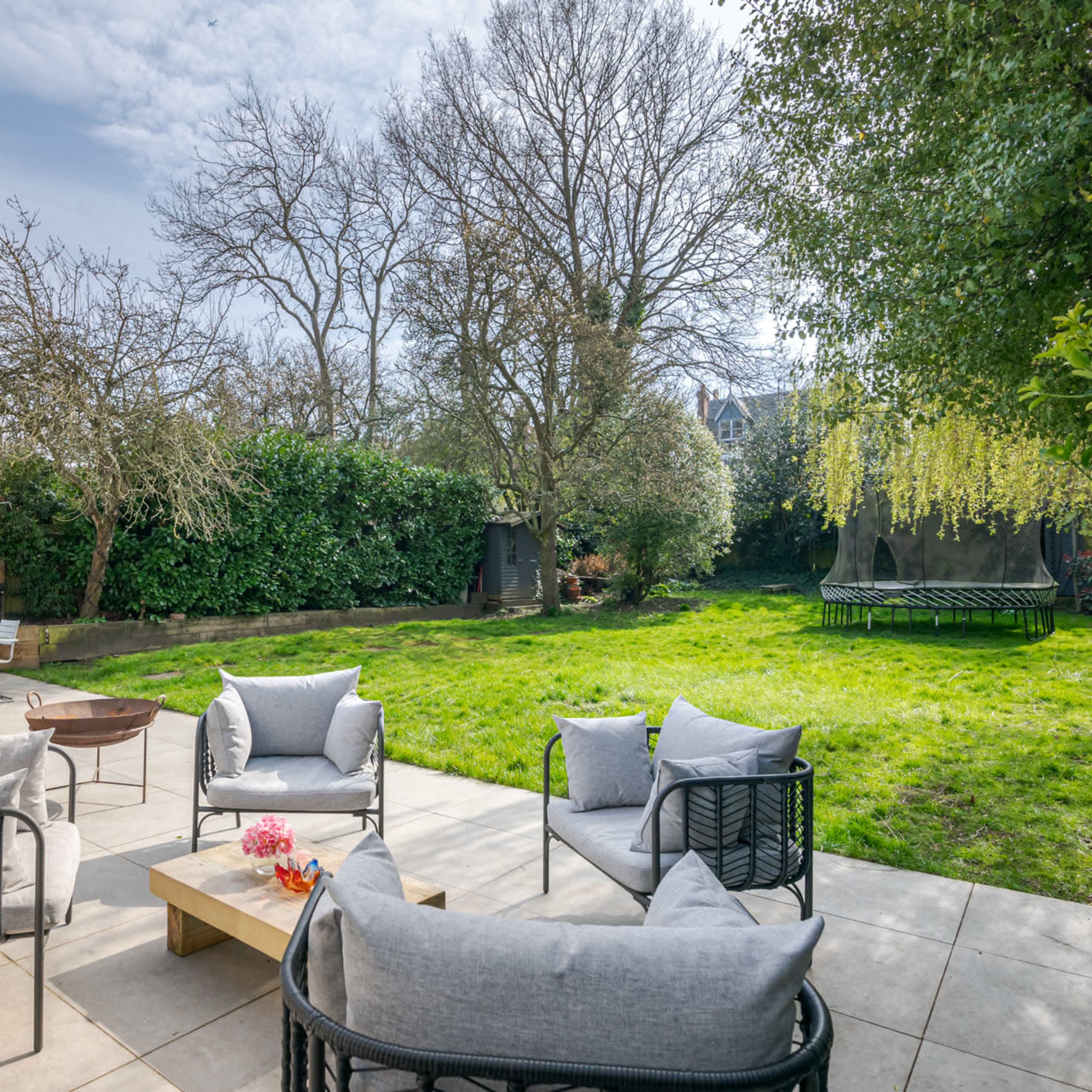 A spacious backyard features a seating area with plush chairs, a wooden coffee table, and a dining table surrounded by trees and greenery.