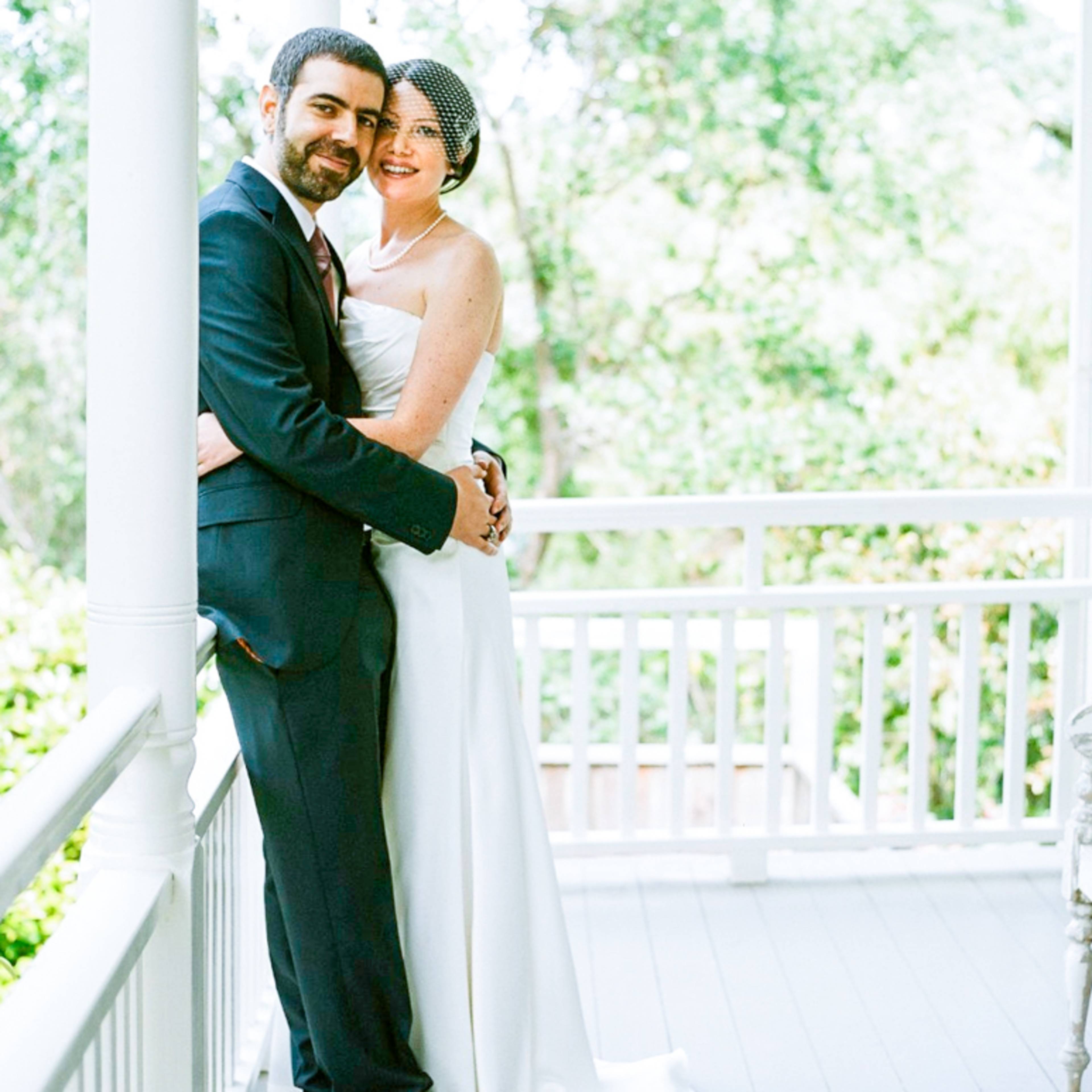 A bride and groom stand together on a porch, embracing each other with greenery visible in the background.