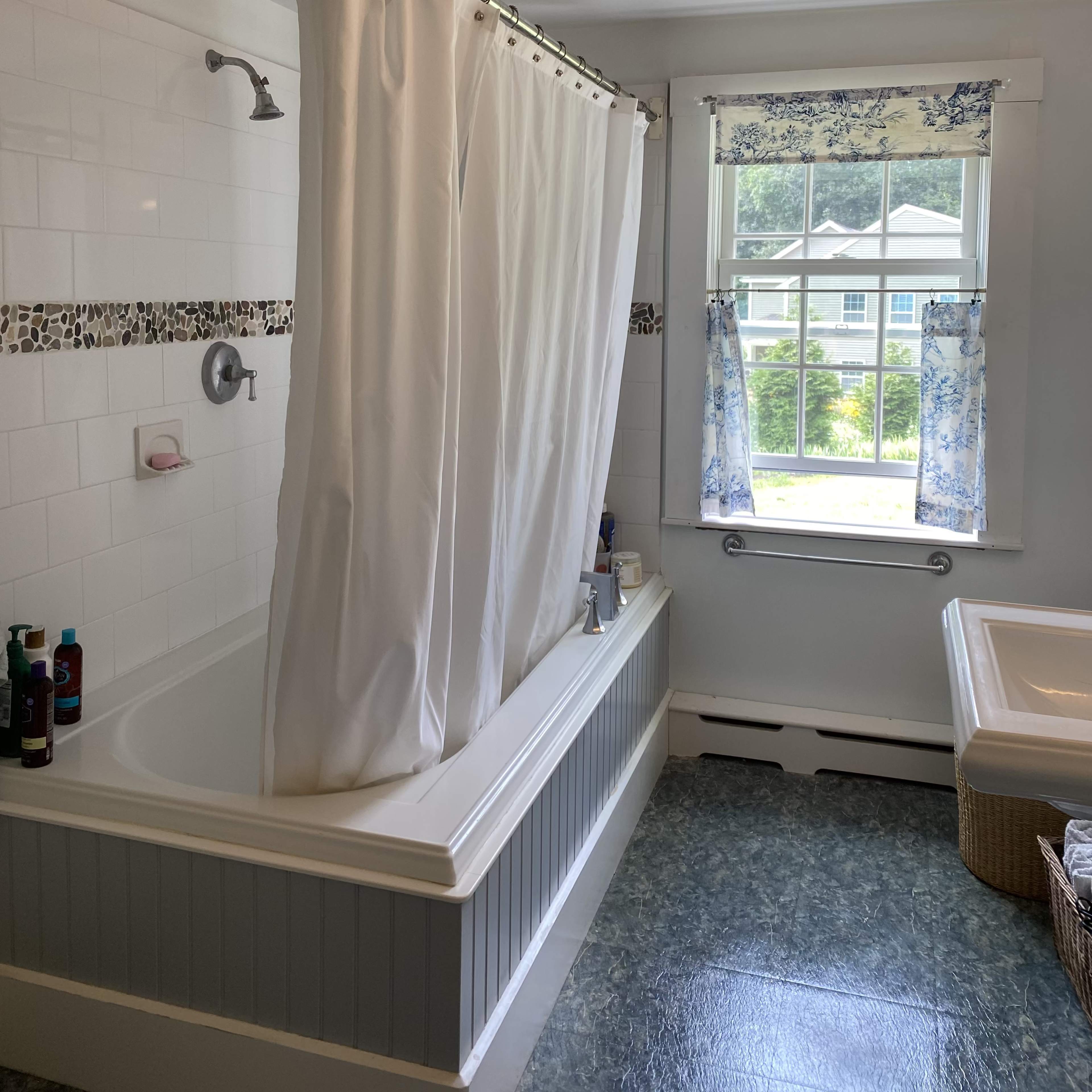 A bathroom with a bathtub and shower combination, a window with patterned curtains, and various toiletries on the ledge.