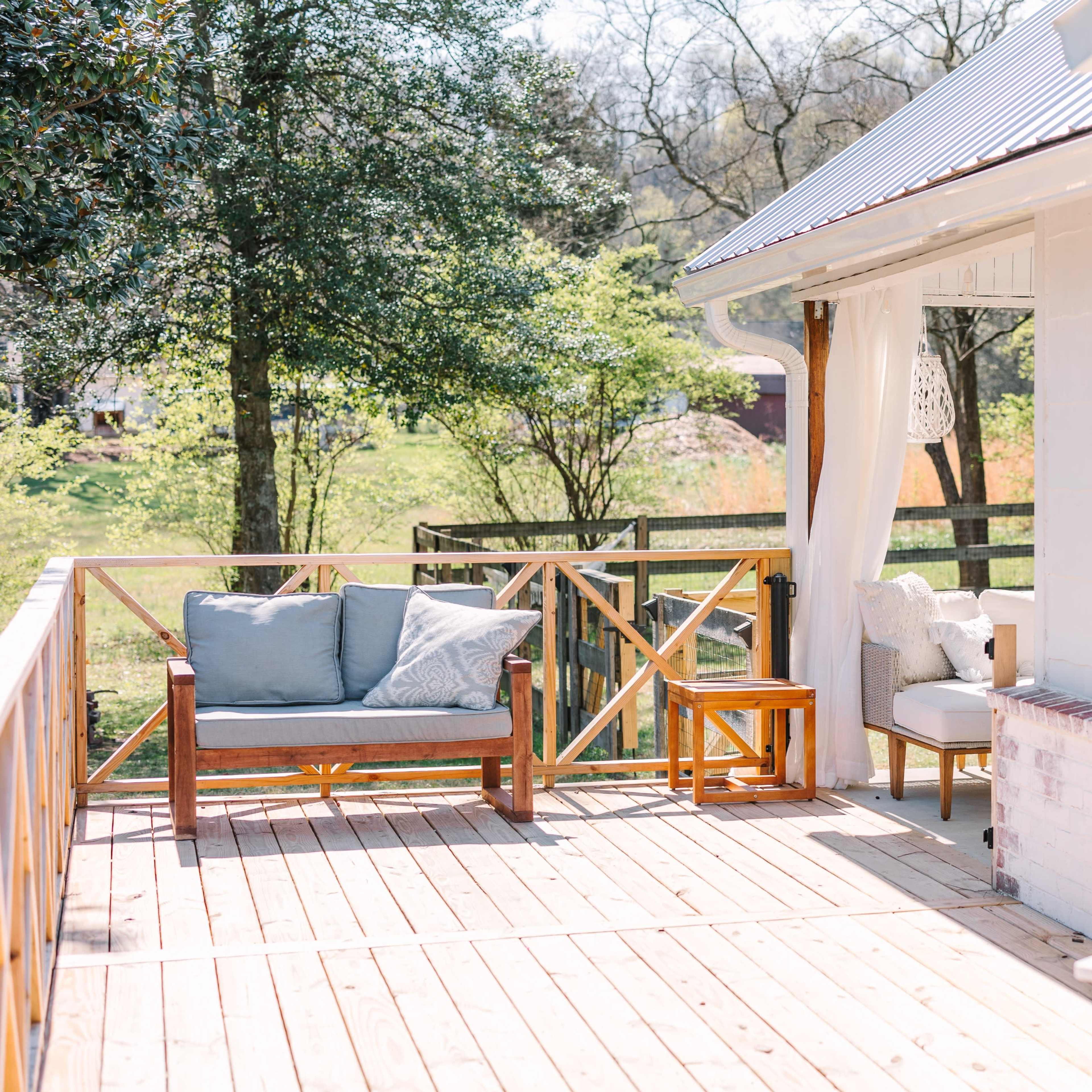 A wooden deck with casual outdoor seating, including a sofa and a side table, surrounded by greenery.