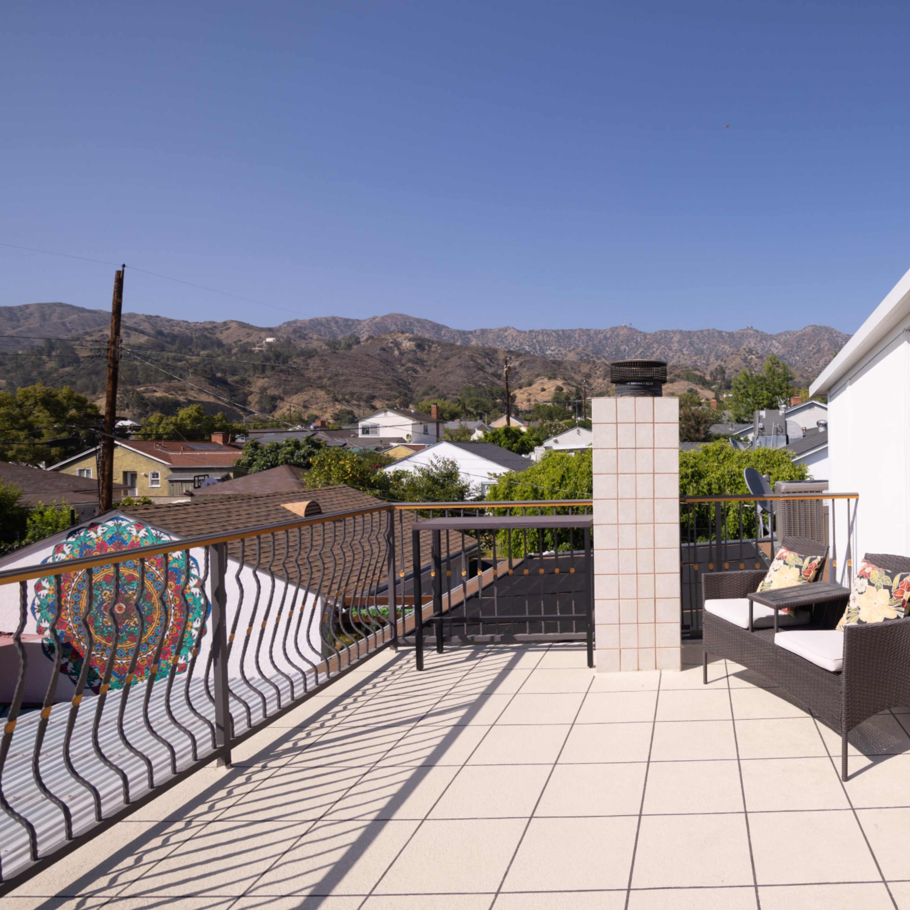 A rooftop terrace with a seating area, a chimney, and a mountainous backdrop under a clear blue sky.