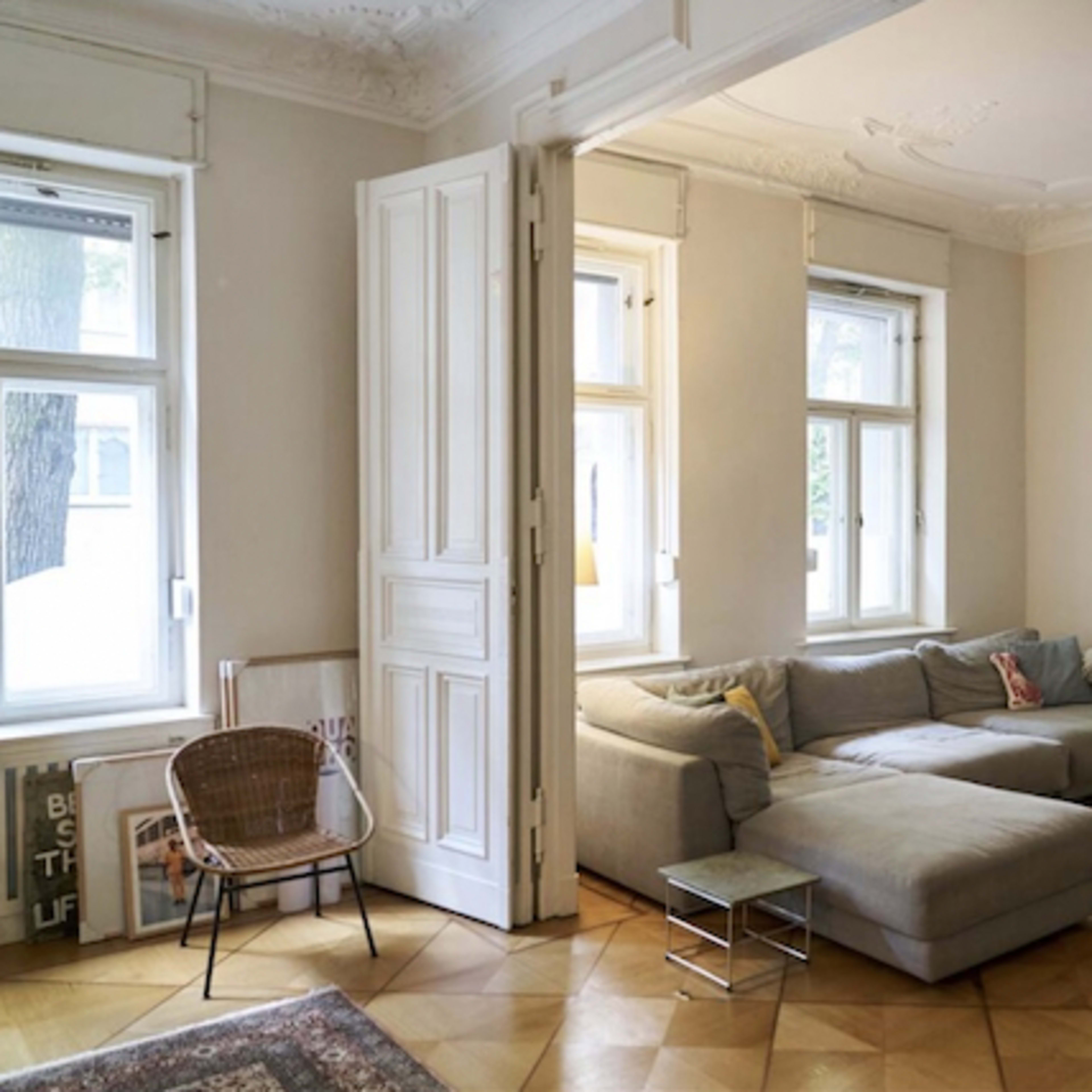 A spacious living room features a gray sectional sofa, a small metal side table, and a wicker chair beside large windows that allow natural light to fill the space.