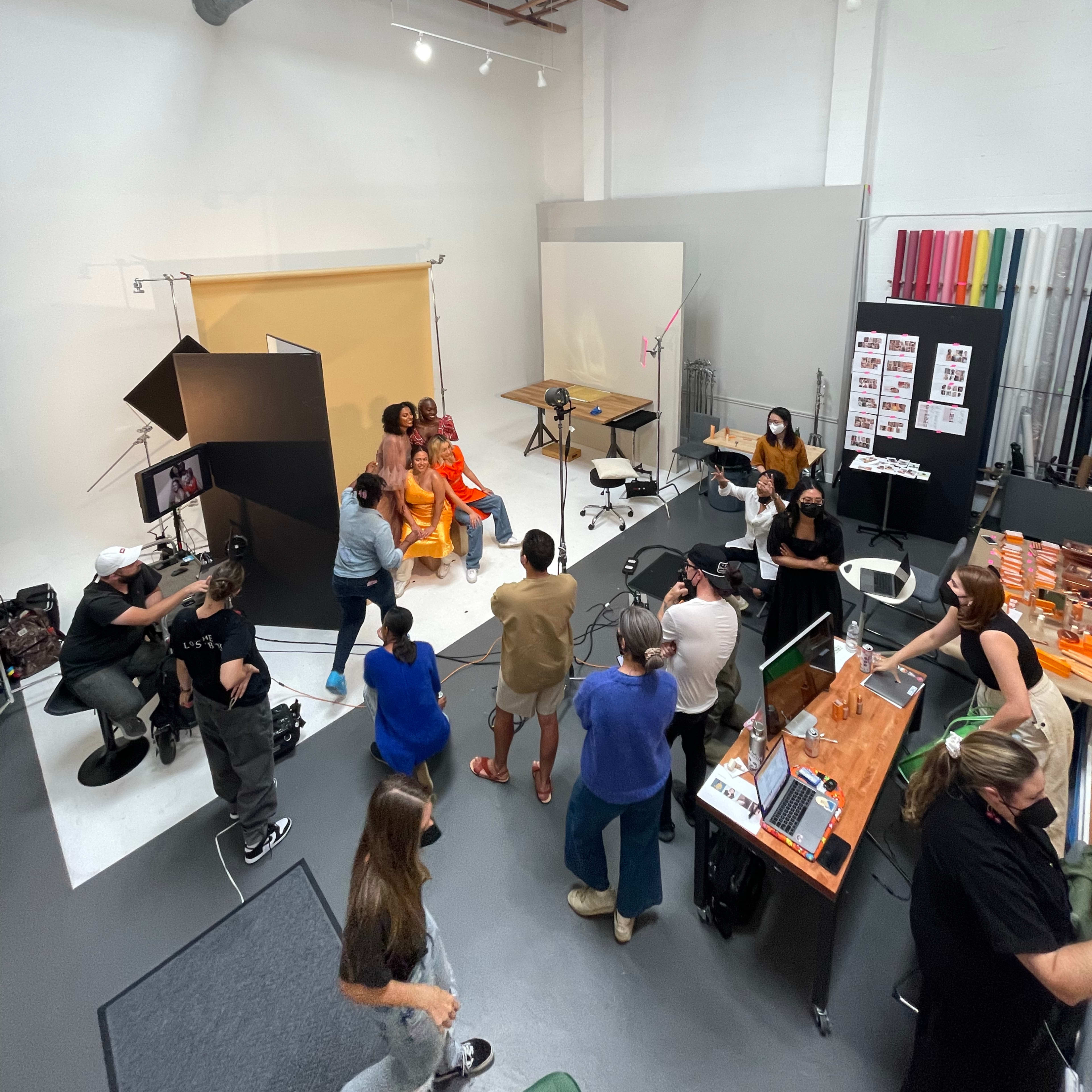 A group of people is gathered in a photography studio, with some arranging equipment and others posing for a photoshoot against a backdrop.