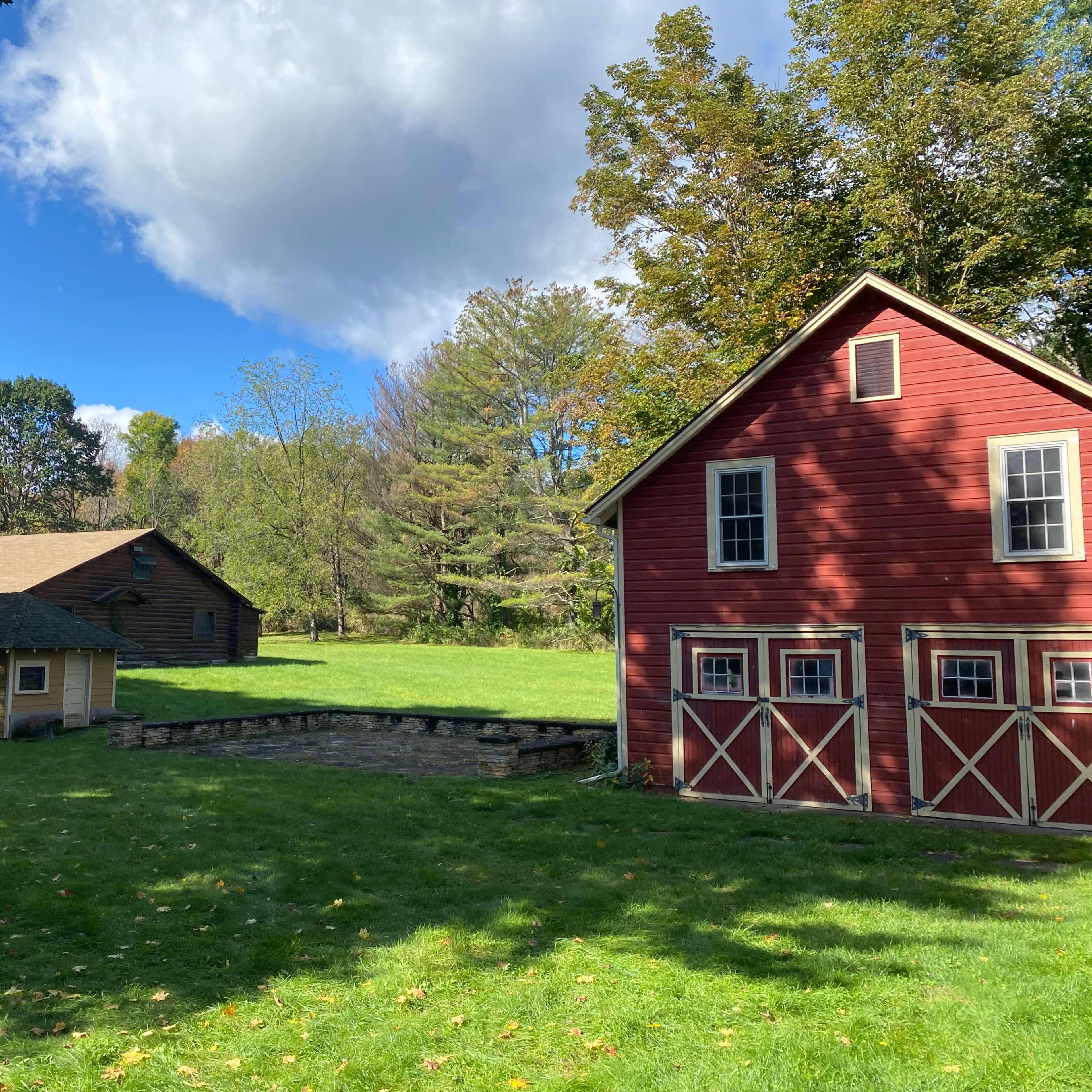 A red house with white trim next to a smaller building and a larger barn, all situated in a grassy area surrounded by trees under a partly cloudy sky.