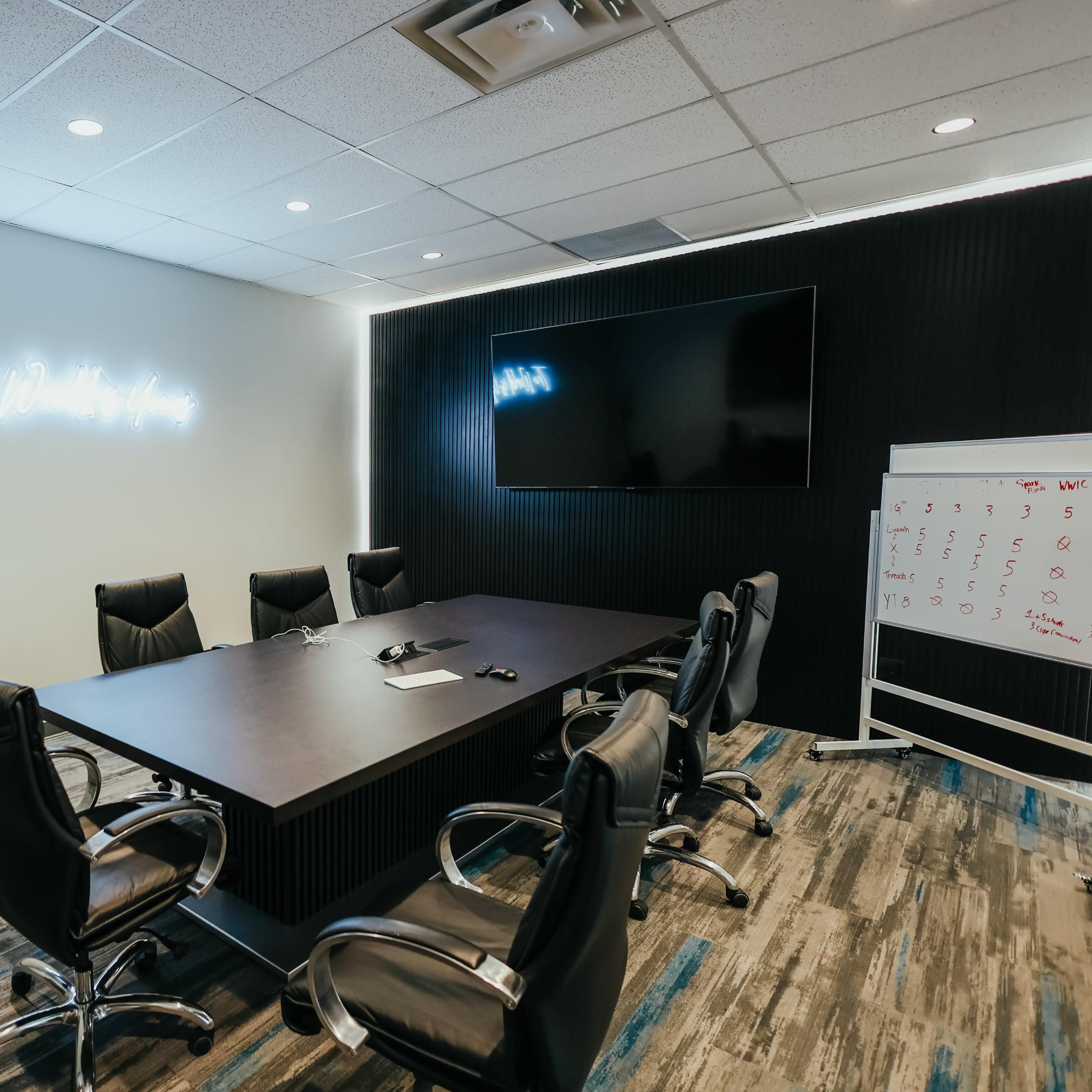 A modern conference room features a large table with eight chairs, a television mounted on the wall, and a whiteboard beside it, all against a backdrop of sleek wall decor and carpeting.