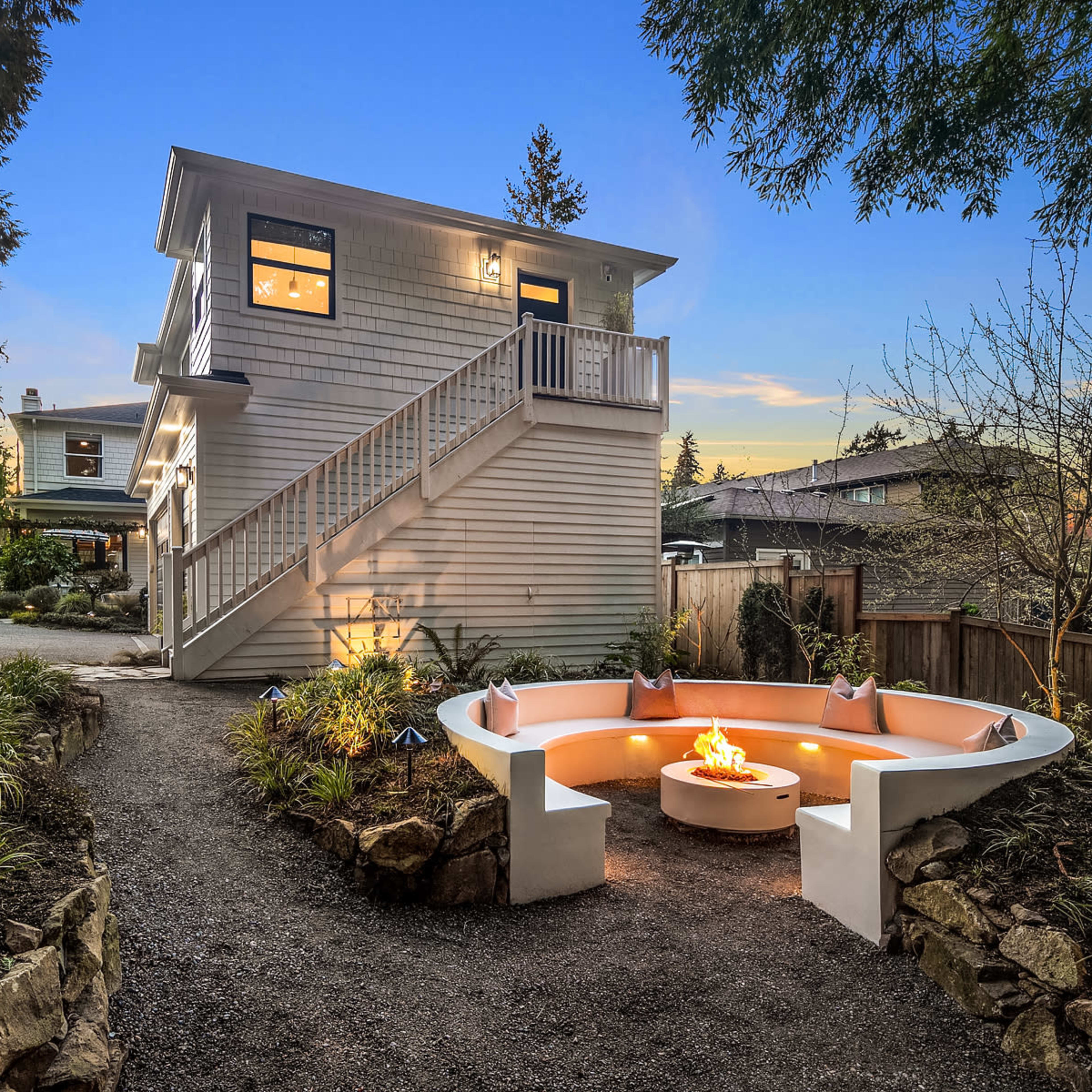 A two-story house with a deck and a fire pit surrounded by landscaped gravel paths and plants.
