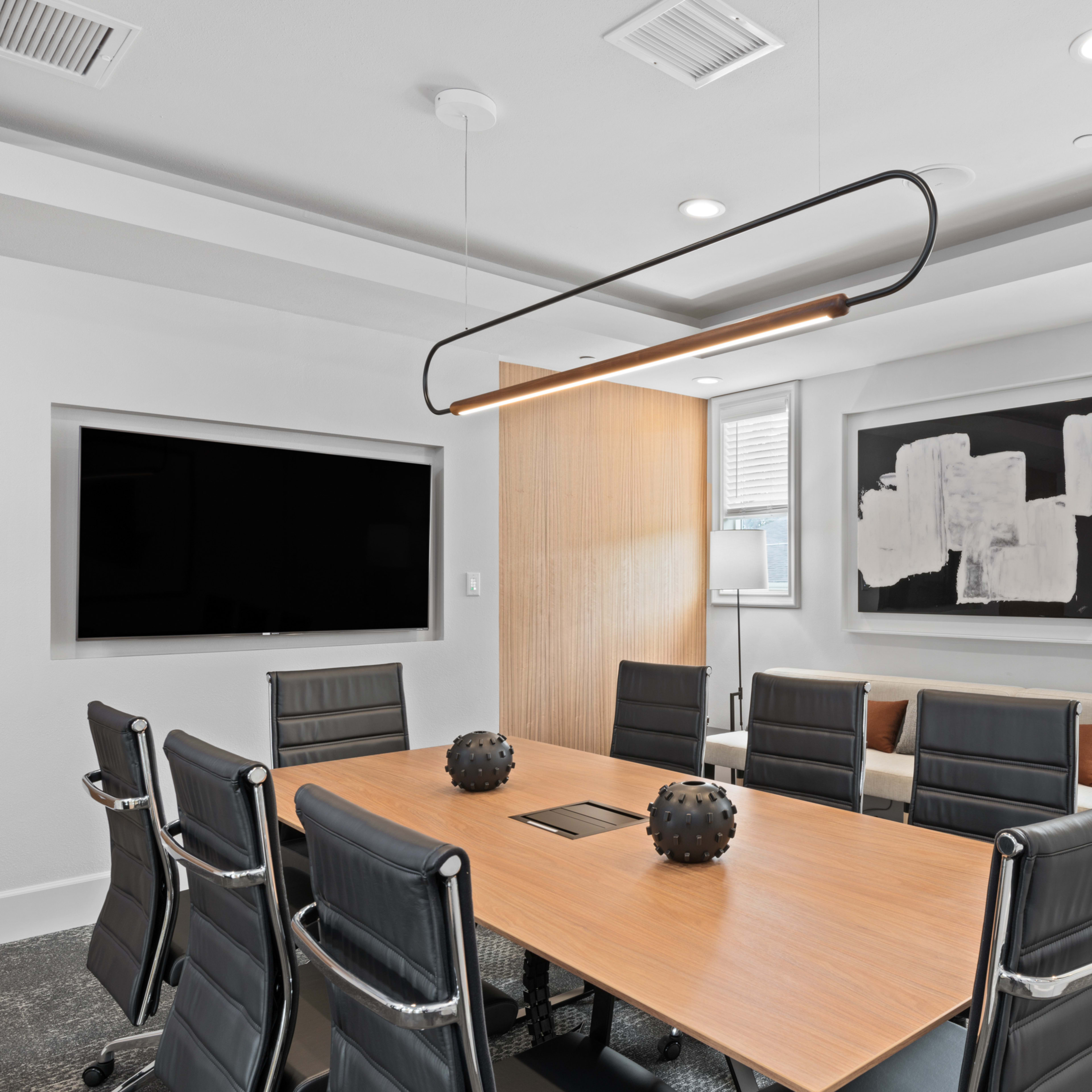 A modern conference room featuring a long wooden table surrounded by black leather chairs, a large screen on the wall, and a minimalist artwork.