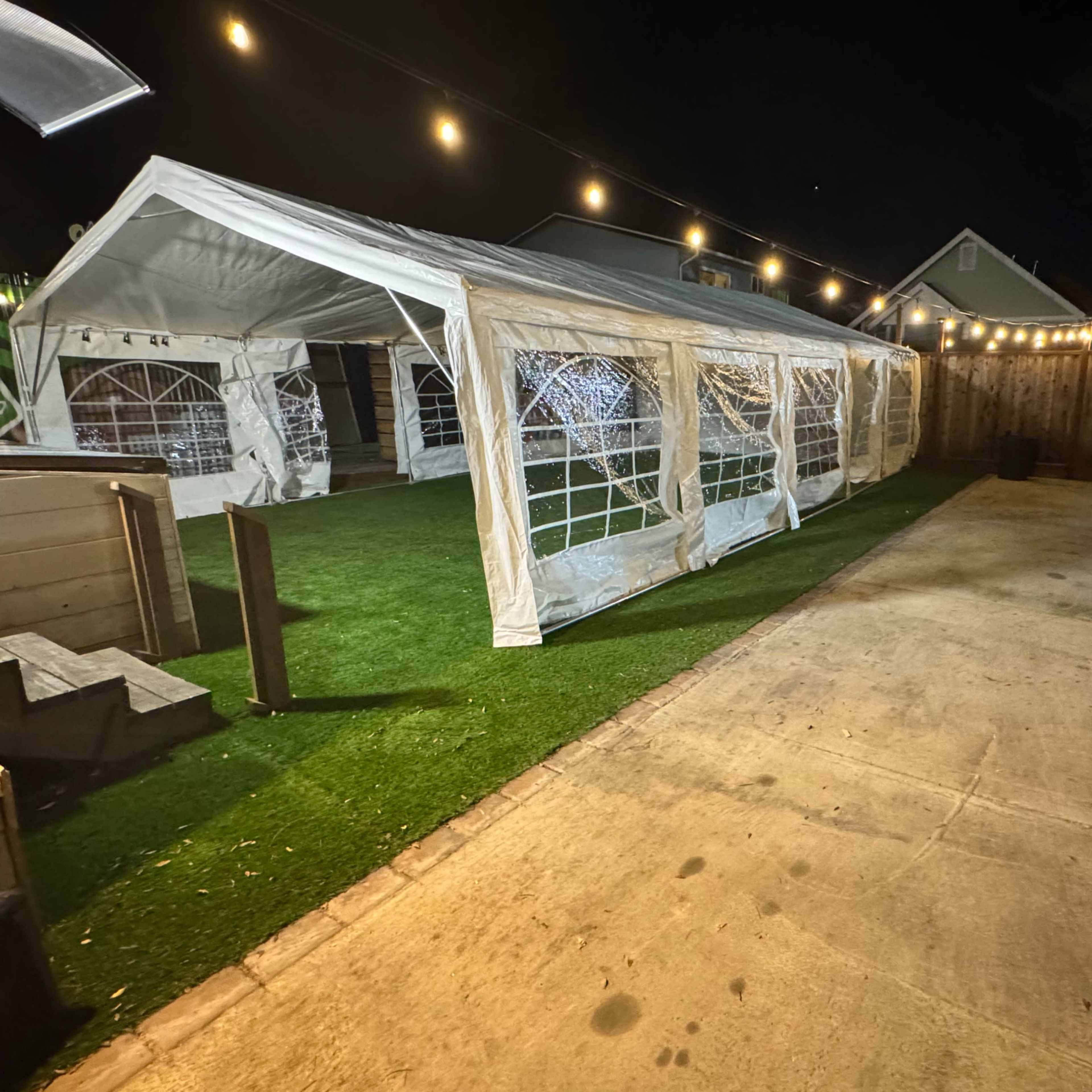 A white tent is set up on a grassy area in a backyard at night, with string lights illuminating the scene.