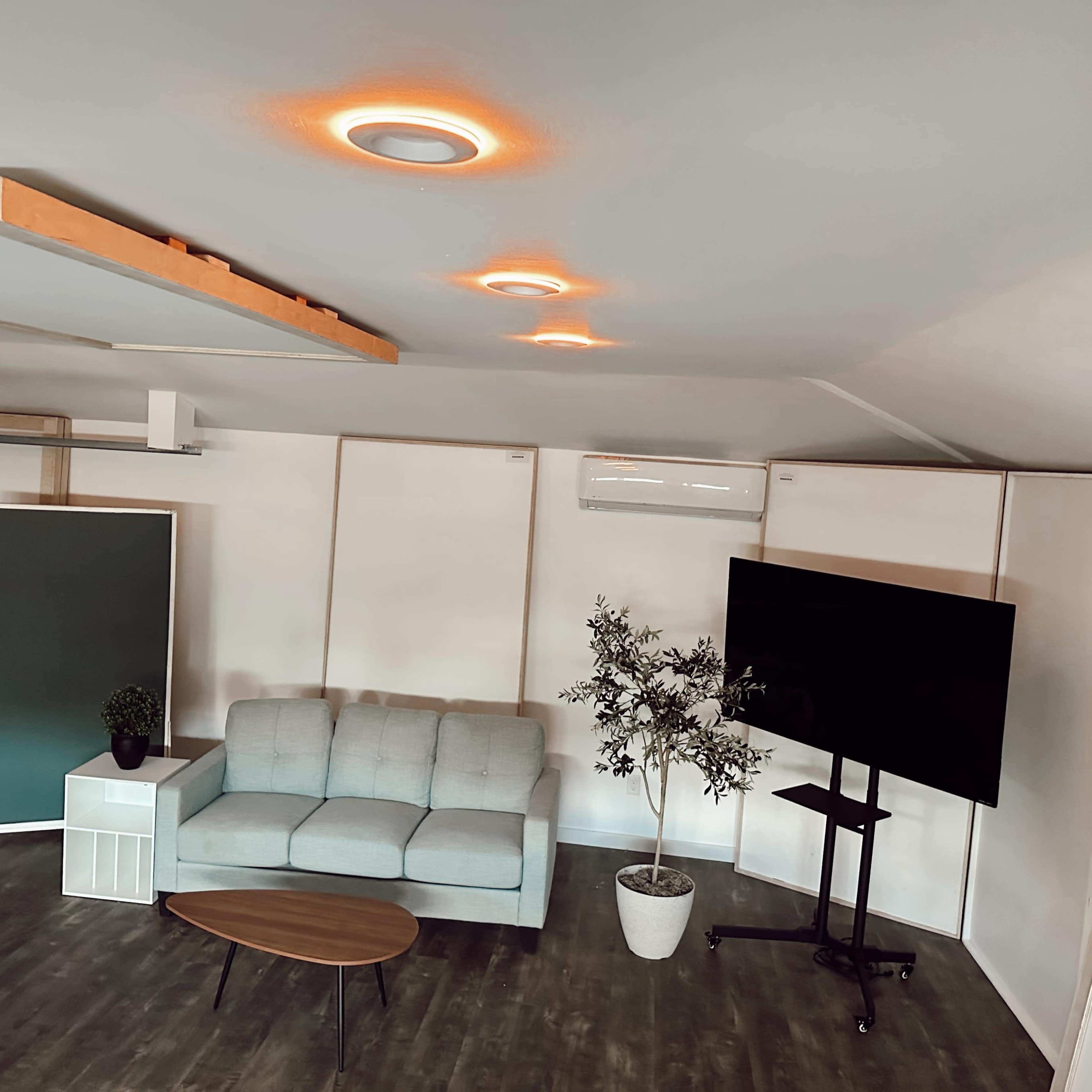 A modern, minimalistic room featuring a gray sofa, a wooden coffee table, a large television on a stand, and a potted plant, all set against a backdrop of white walls and a wooden ceiling.